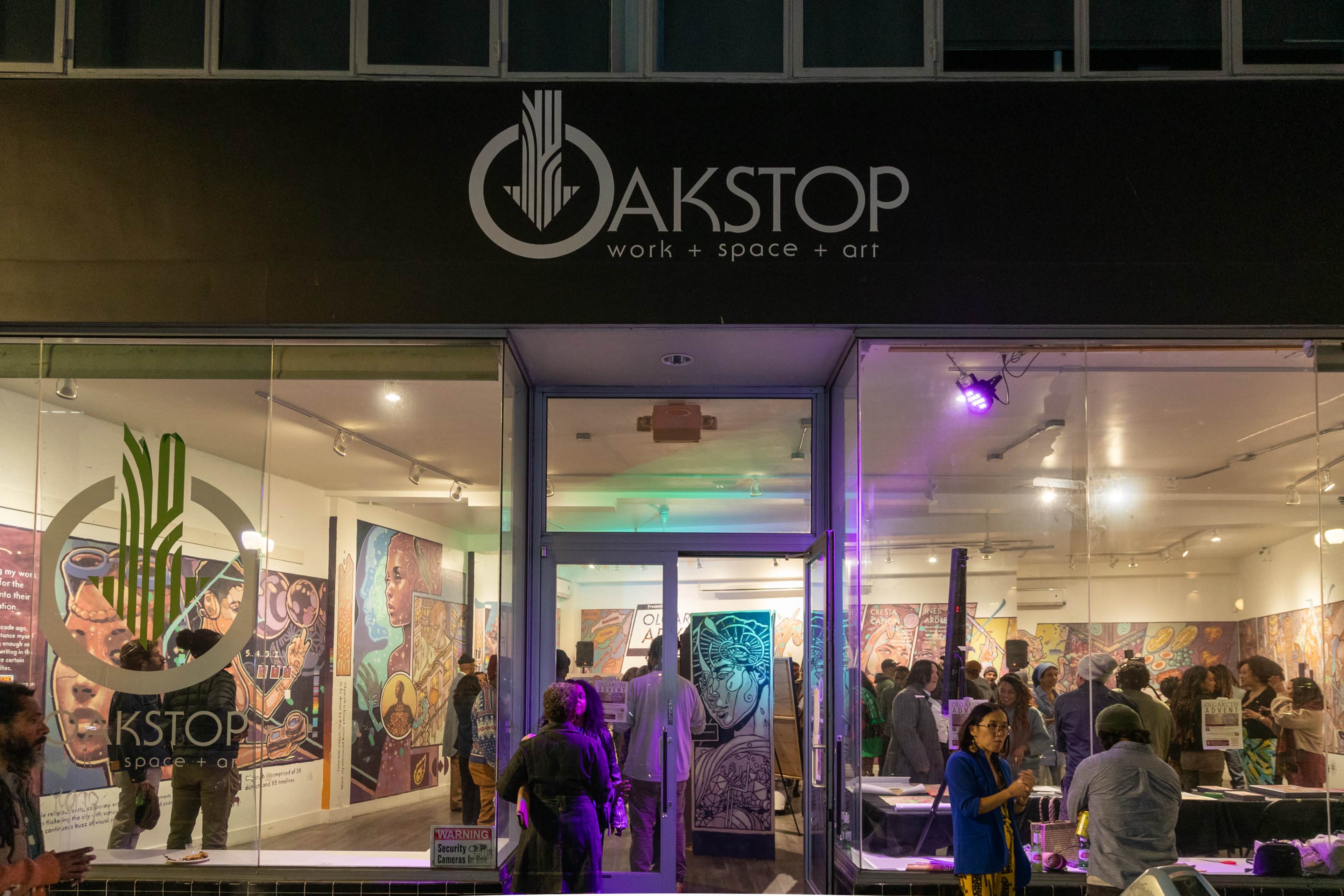 The image shows a modern gallery space named "Oakstop," where people are gathered, admiring artwork displayed on the walls.