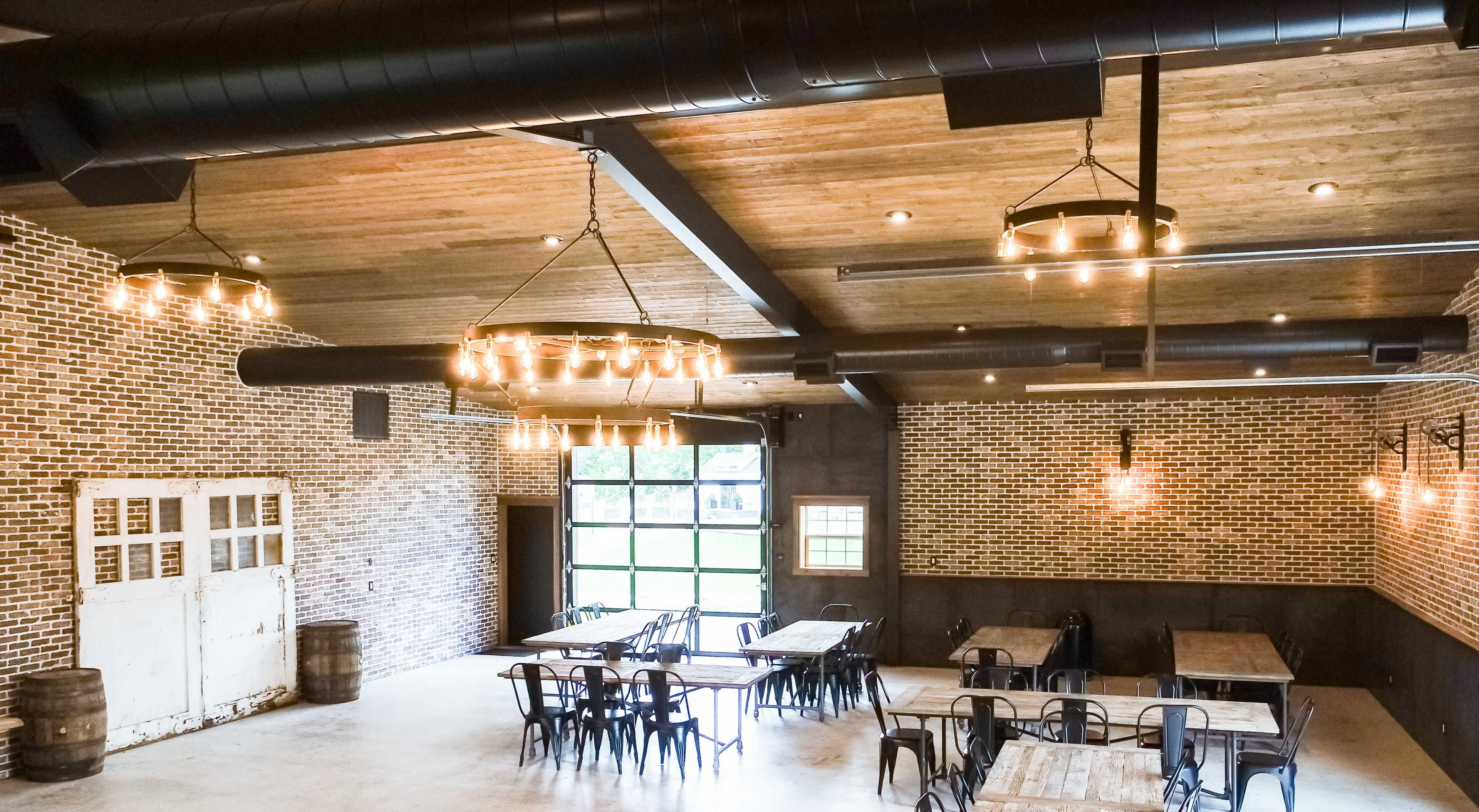 The image shows a spacious event venue with exposed brick walls, wooden beams, and several tables arranged beneath industrial-style light fixtures.