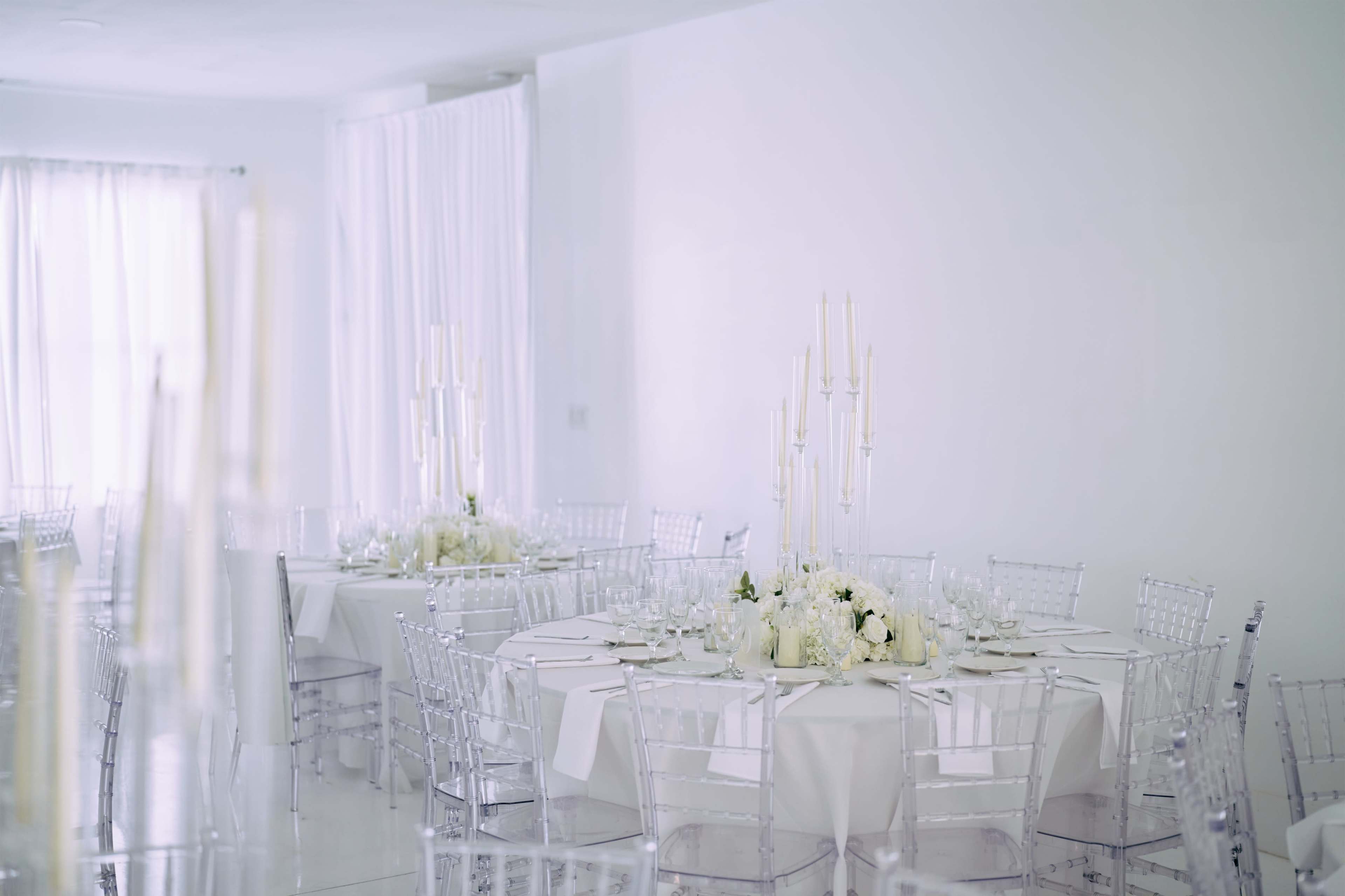 The image shows a minimalist event space with white walls and decorated tables, featuring clear chairs and elegant floral centerpieces.