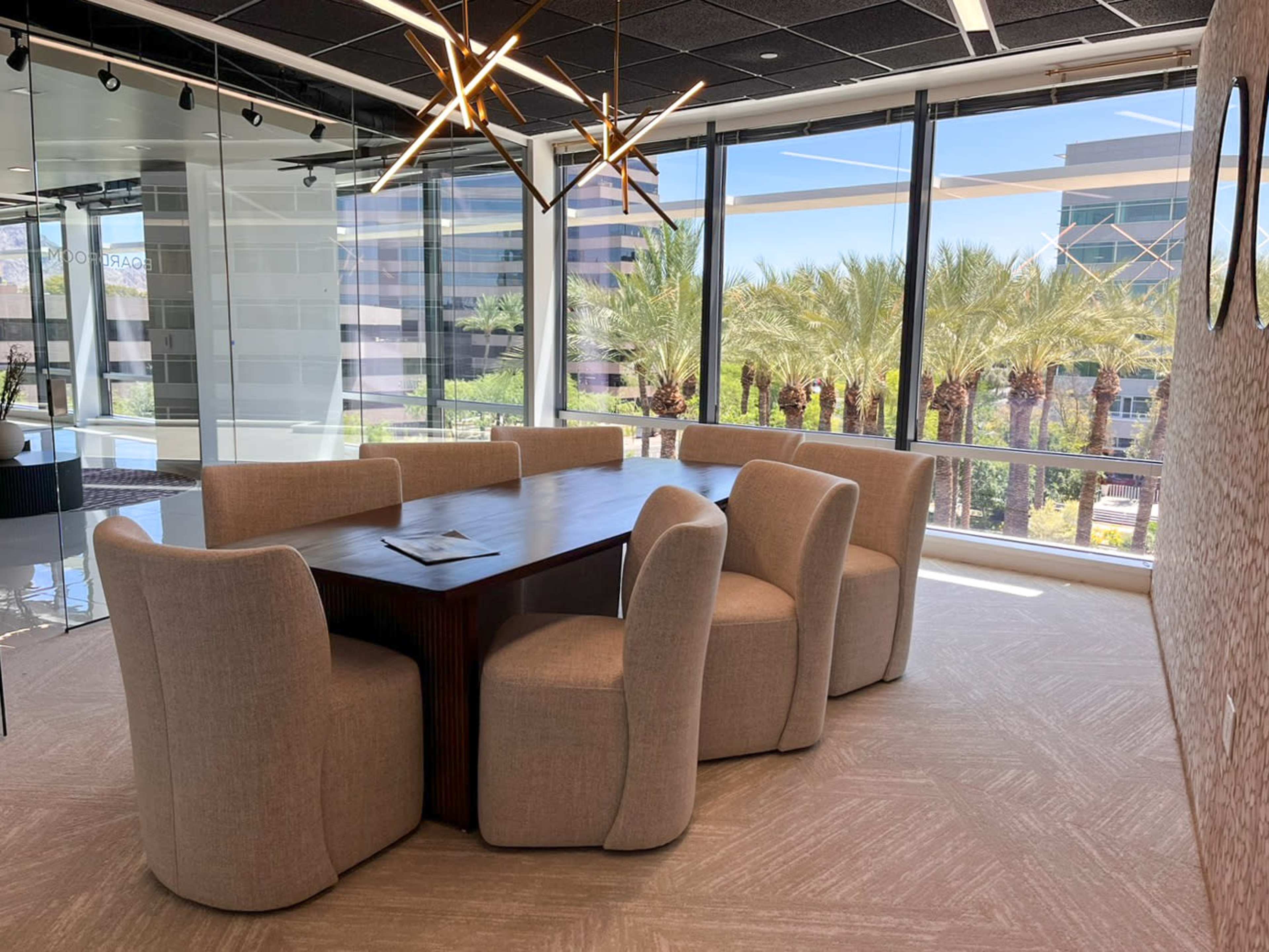 A modern conference room features a large wooden table surrounded by six upholstered chairs, with large windows offering a view of palm trees outside.