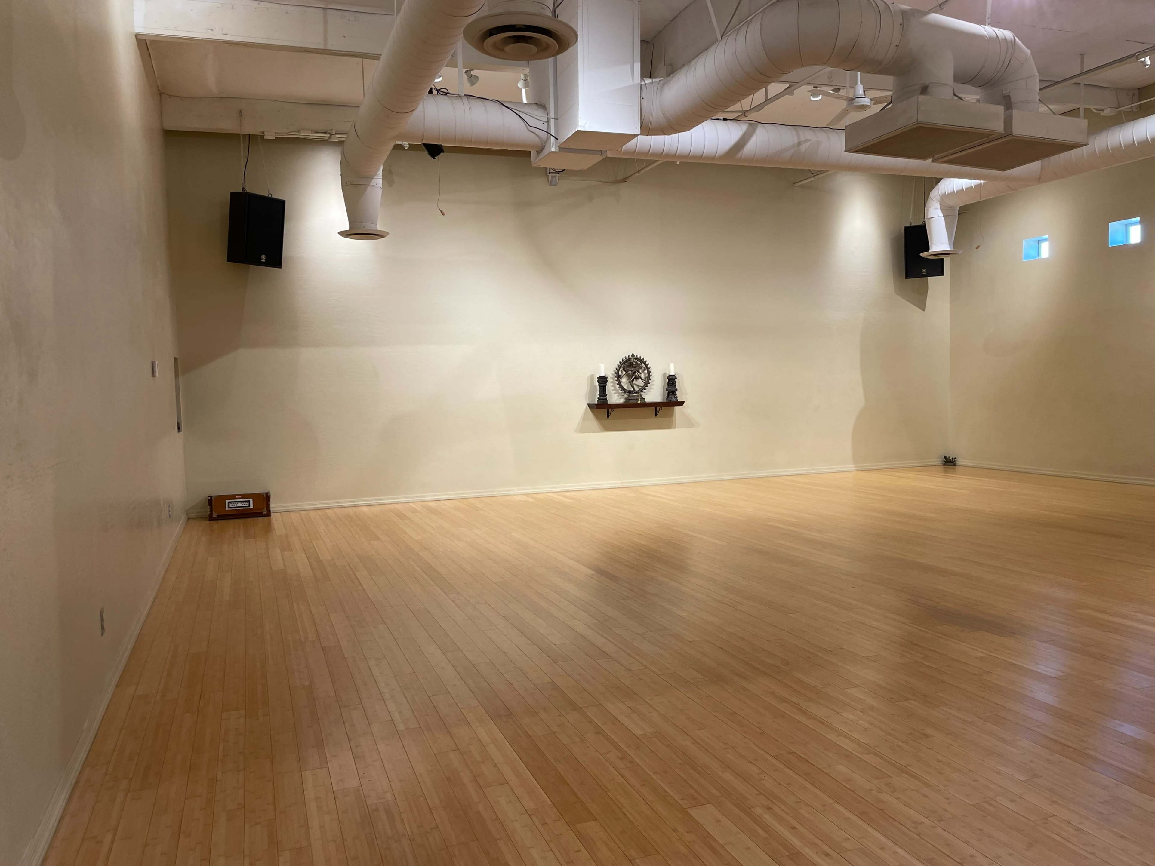 A spacious, empty studio with wooden flooring, beige walls, and a small shelf displaying decorative items.