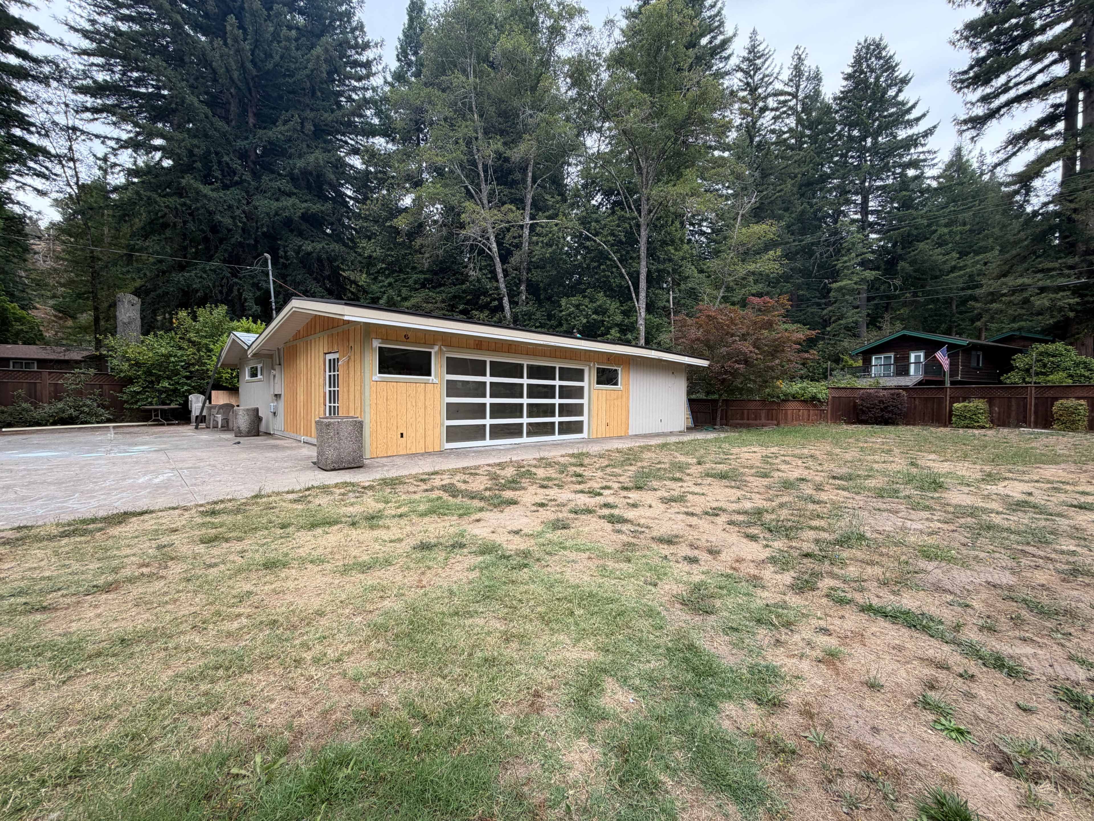 A modern single-story house with a large glass garage door sits on a grassy plot surrounded by tall trees and other homes in the background.