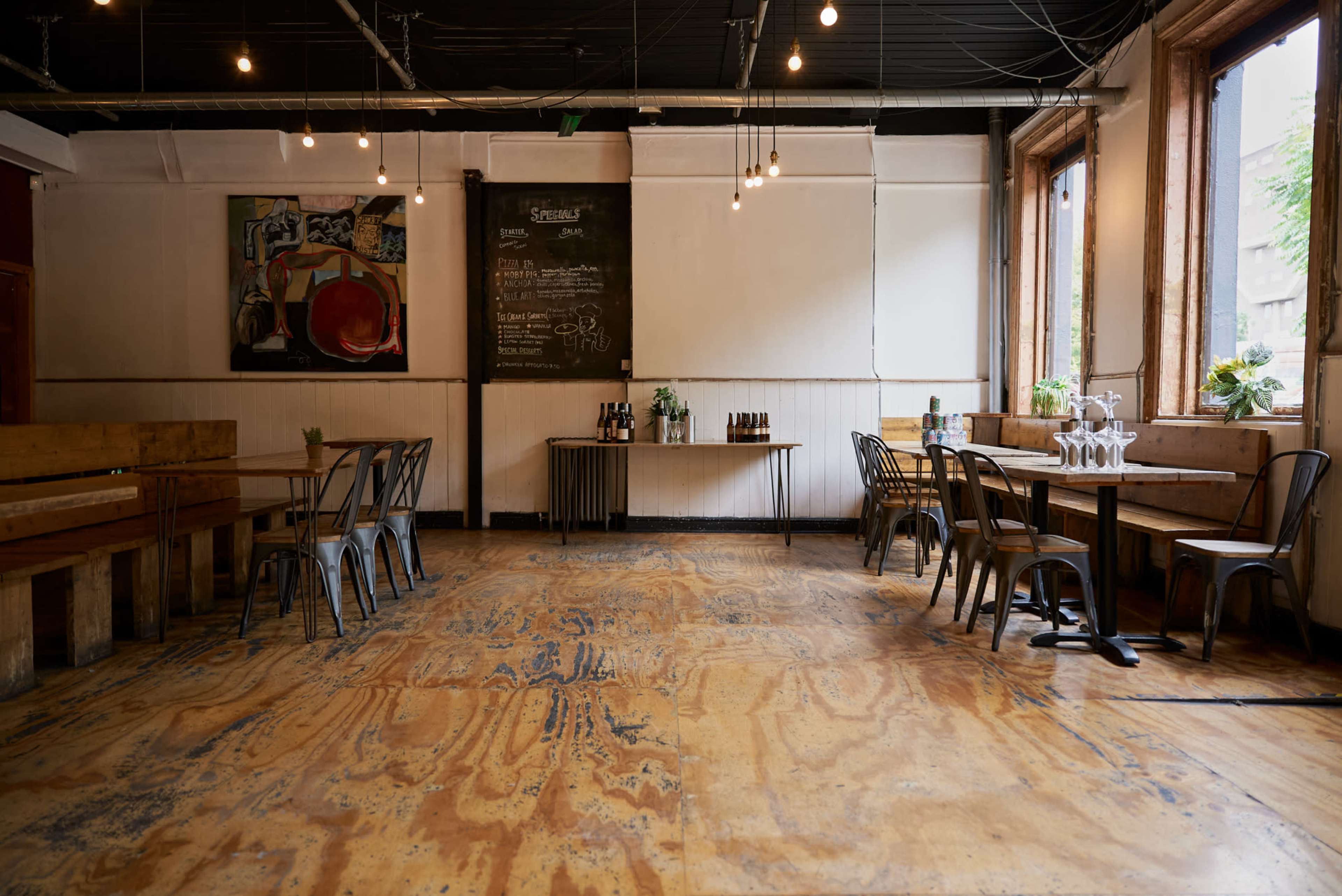 The image shows a spacious, minimalistic restaurant interior with wooden tables, metal chairs, and large windows.