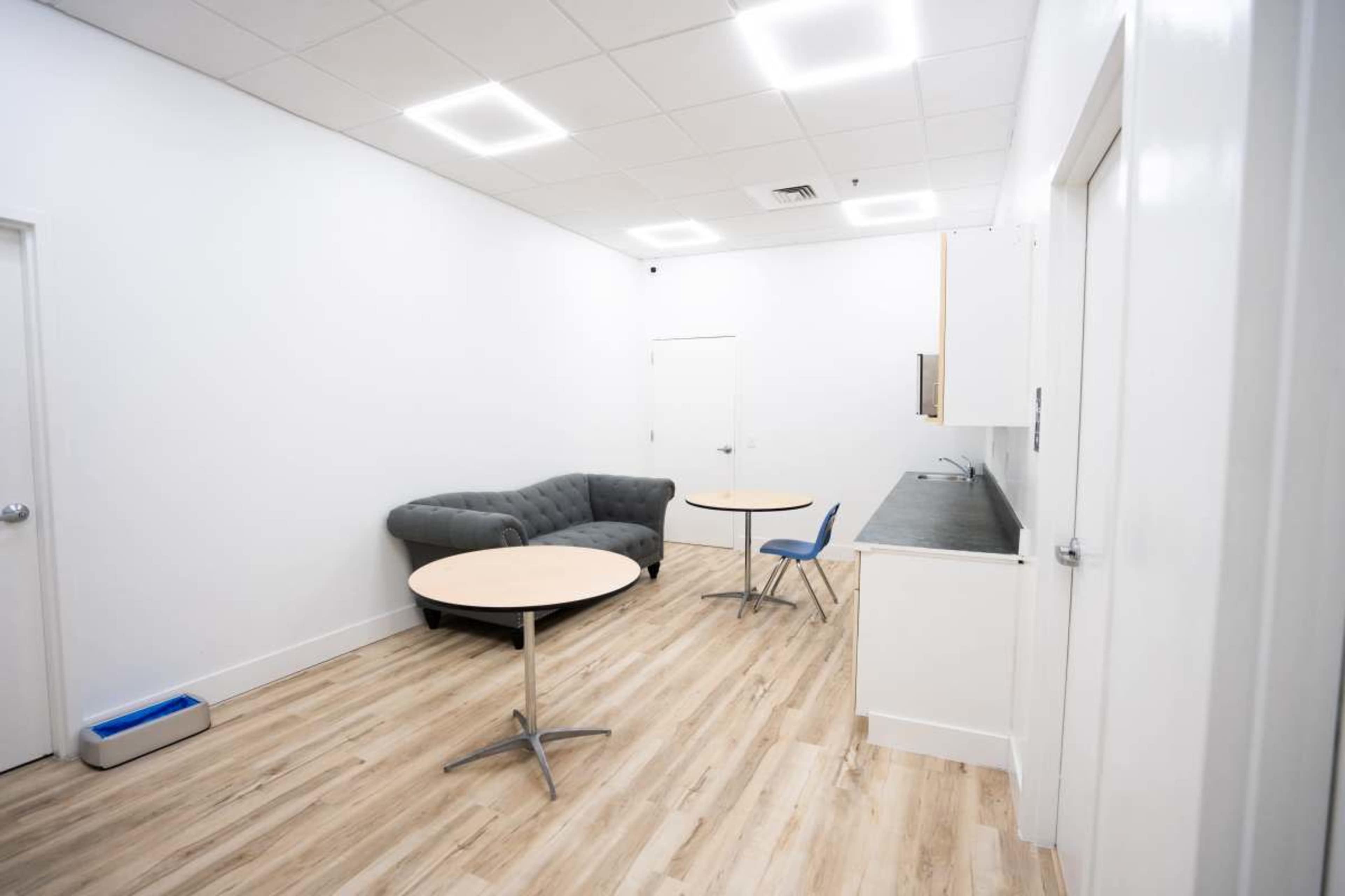 The image shows a small, modern room with two round tables, a gray sofa, and a kitchenette area.