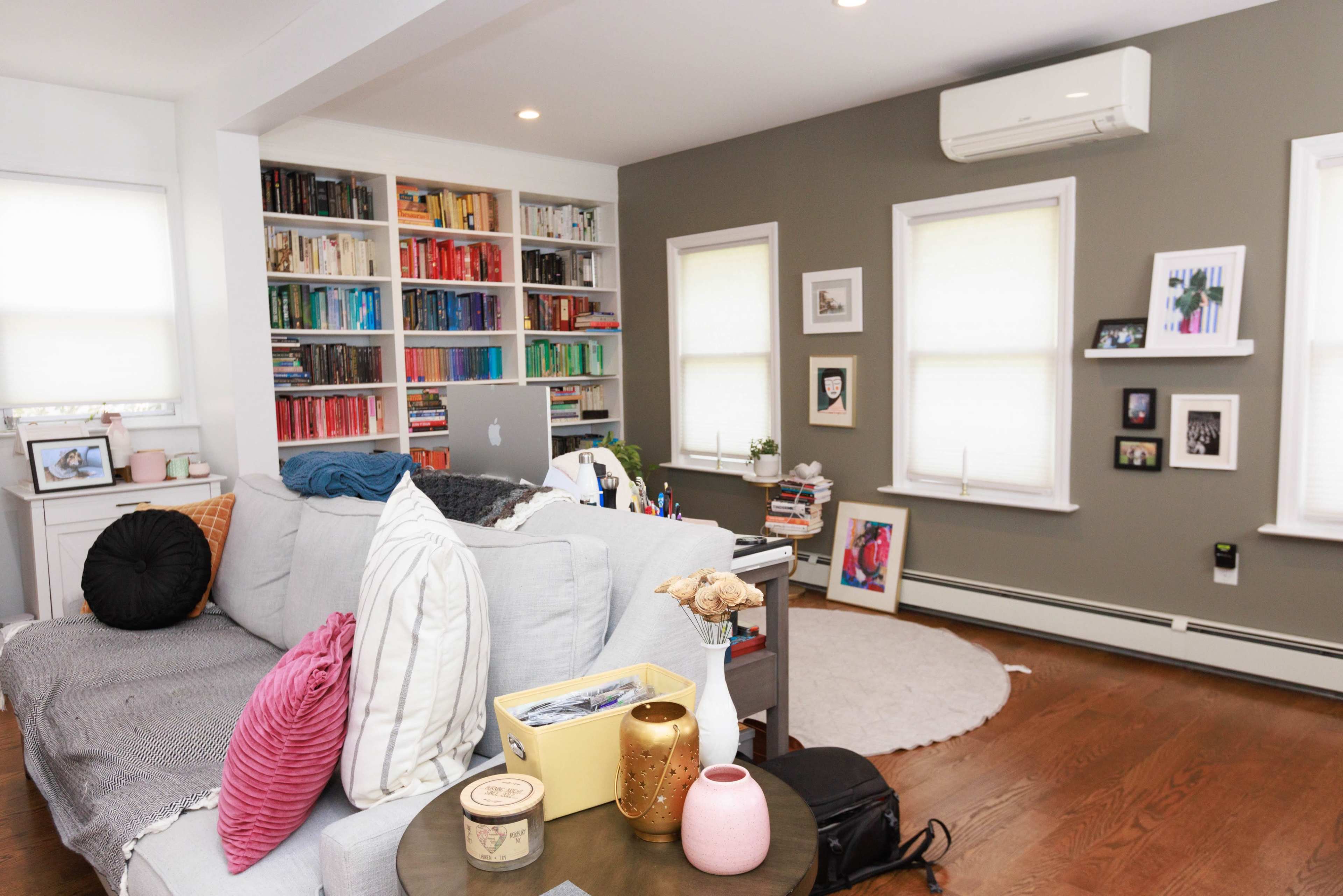 A living room features a light-colored sectional sofa, a circular area rug, a wall of colorful bookshelves, and several windows with blinds.