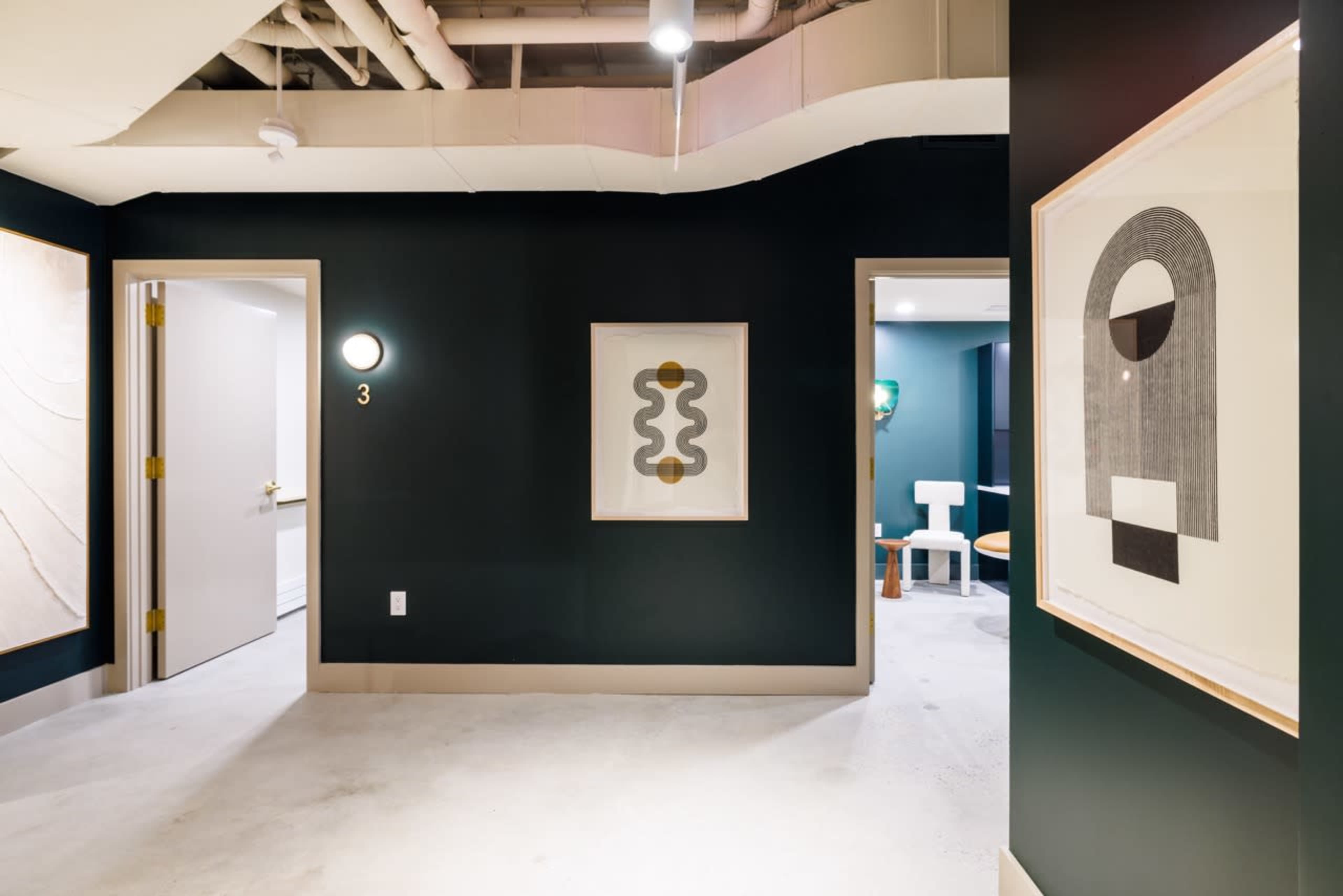 A hallway with dark green walls features framed artwork and a doorway leading to another room.