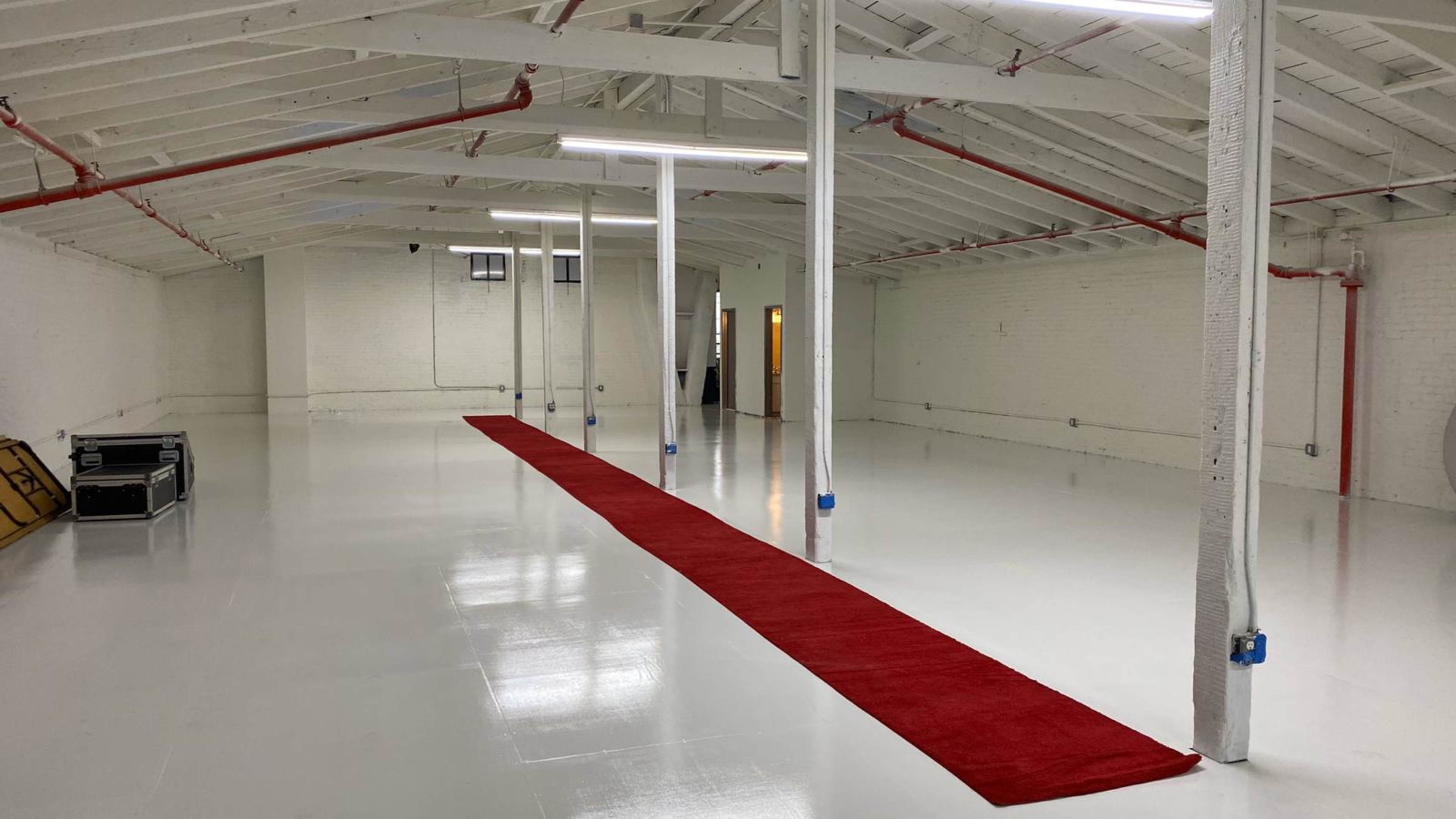 A spacious, empty room with a red carpet running along the center, illuminated by overhead lights and featuring white walls and a high ceiling supported by columns.