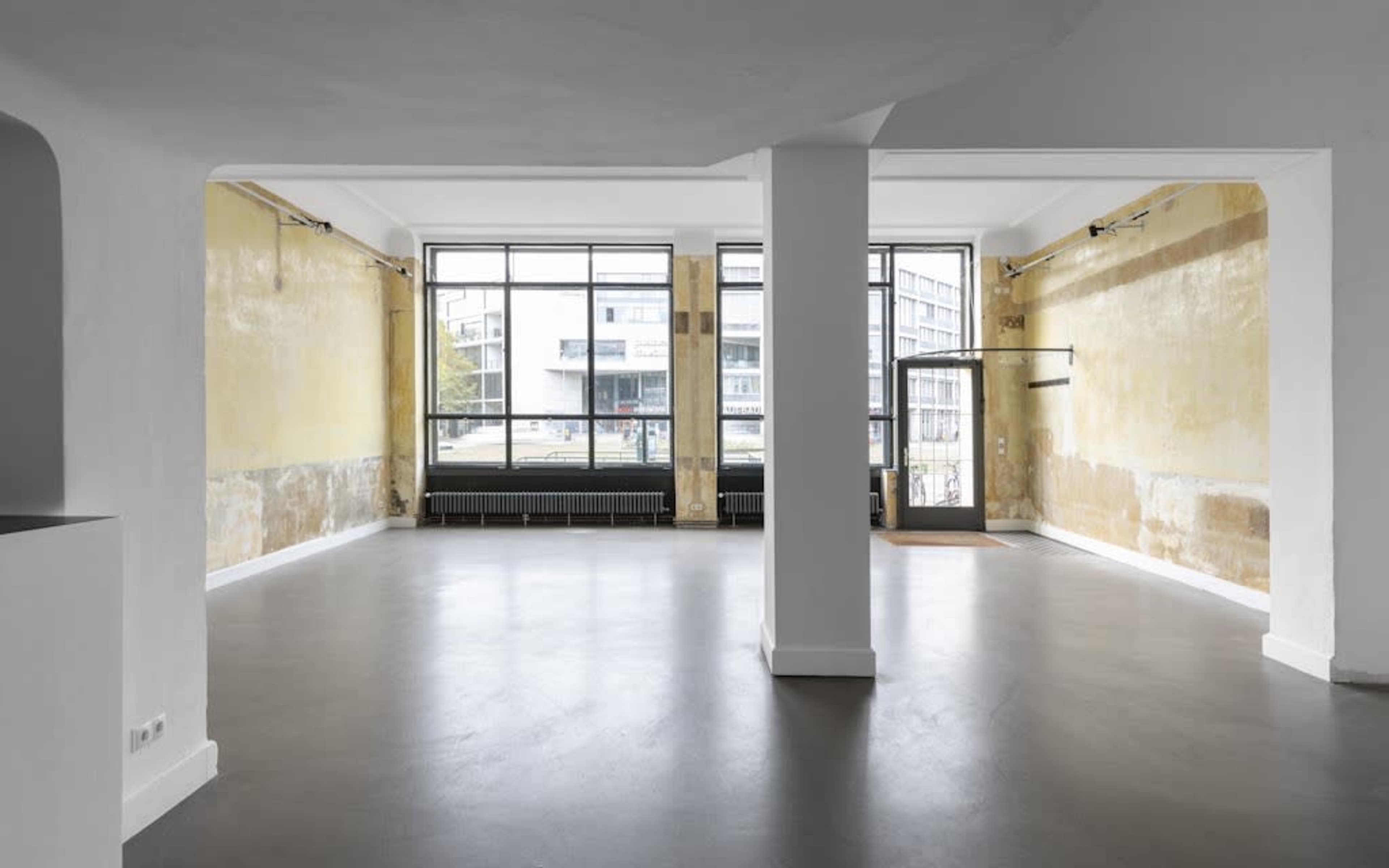 An empty room with large windows, featuring yellow-painted walls, a concrete floor, and a column in the center.