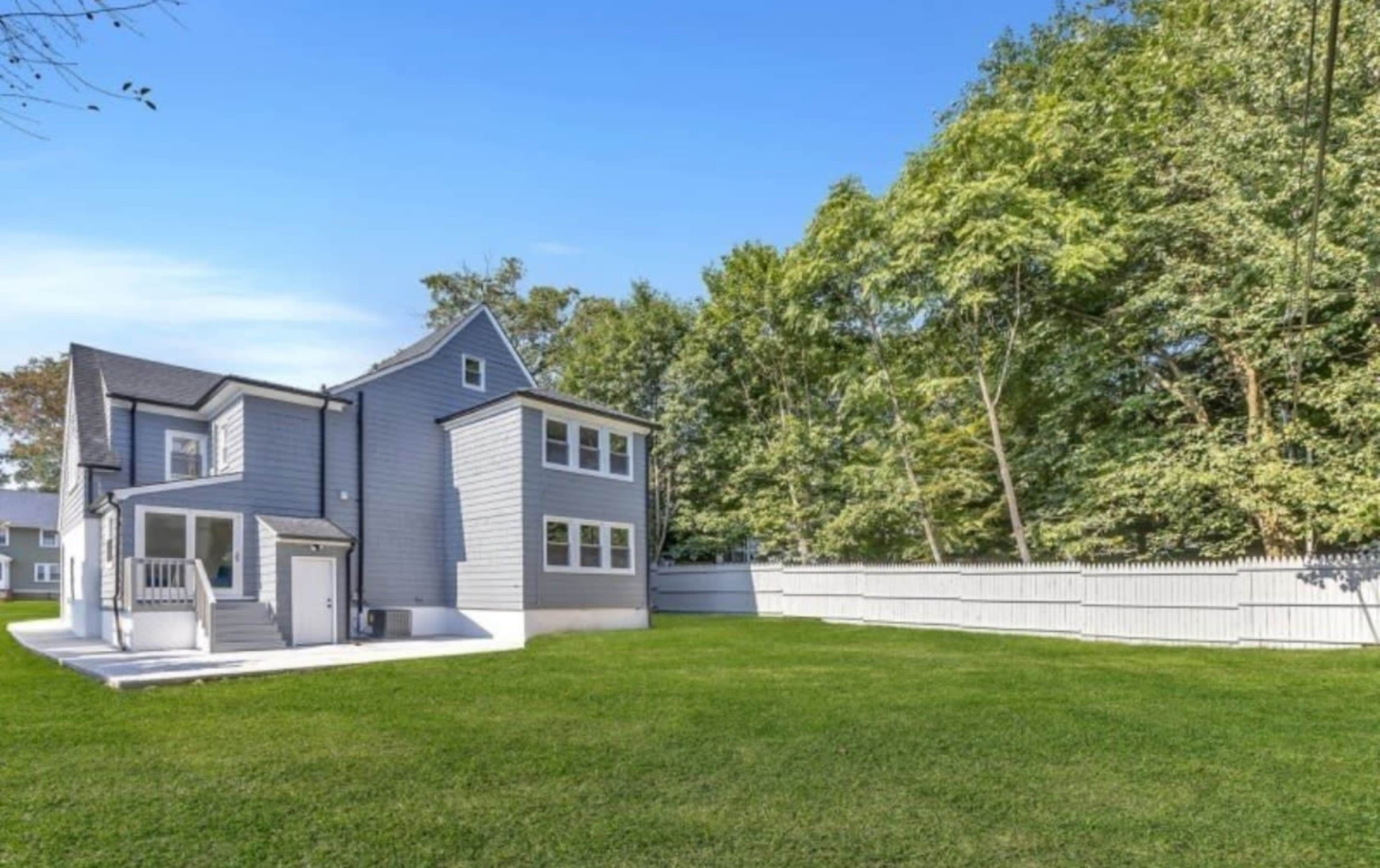 A two-story gray house sits in a spacious backyard surrounded by trees and a white privacy fence.