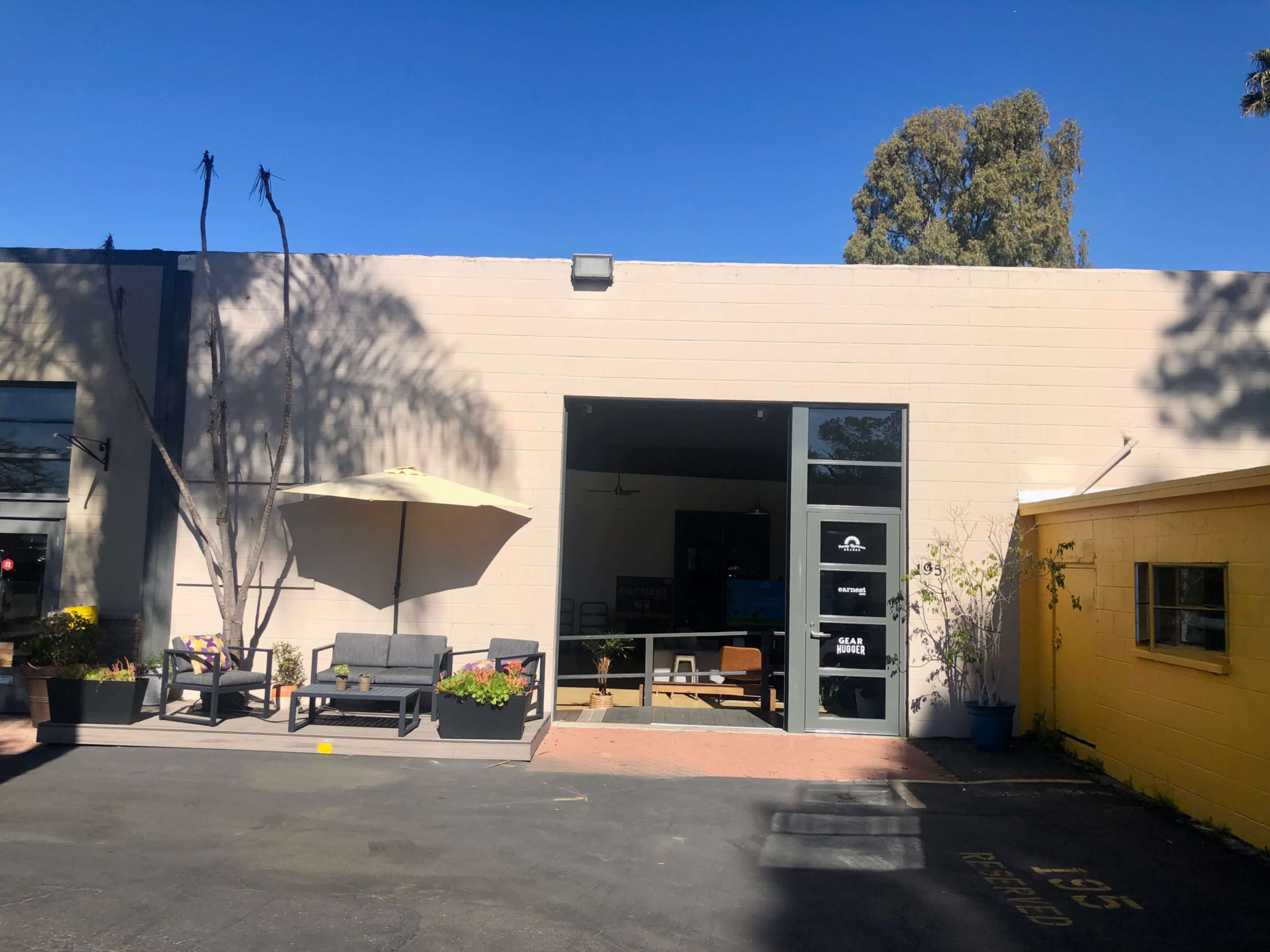 A commercial building with a large open entrance, featuring outdoor seating and an umbrella beside a yellow structure in a paved area.