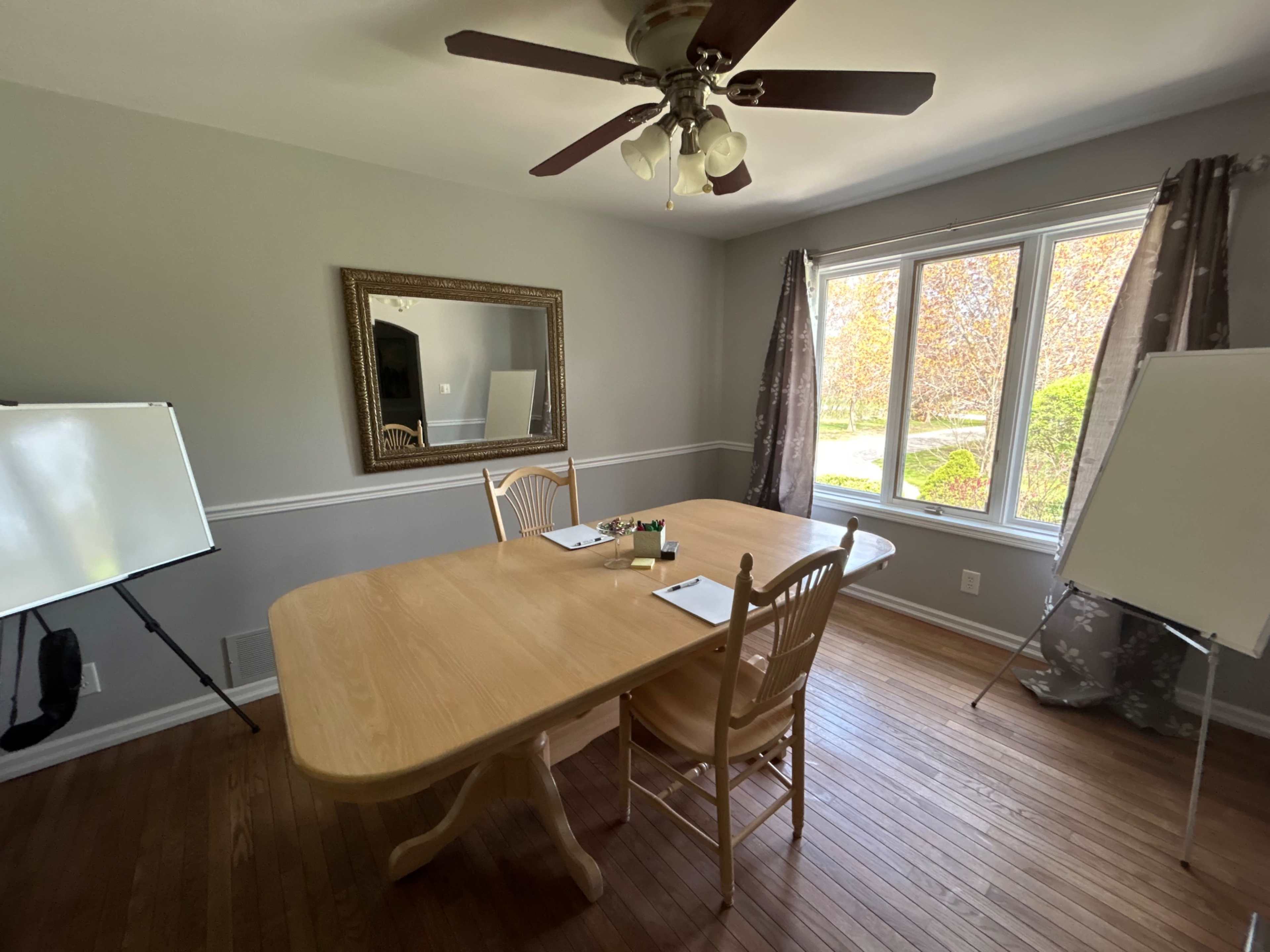 A conference room features a large wooden table with two chairs, a mirror on the wall, and two whiteboards on either side, all illuminated by natural light from the windows.
