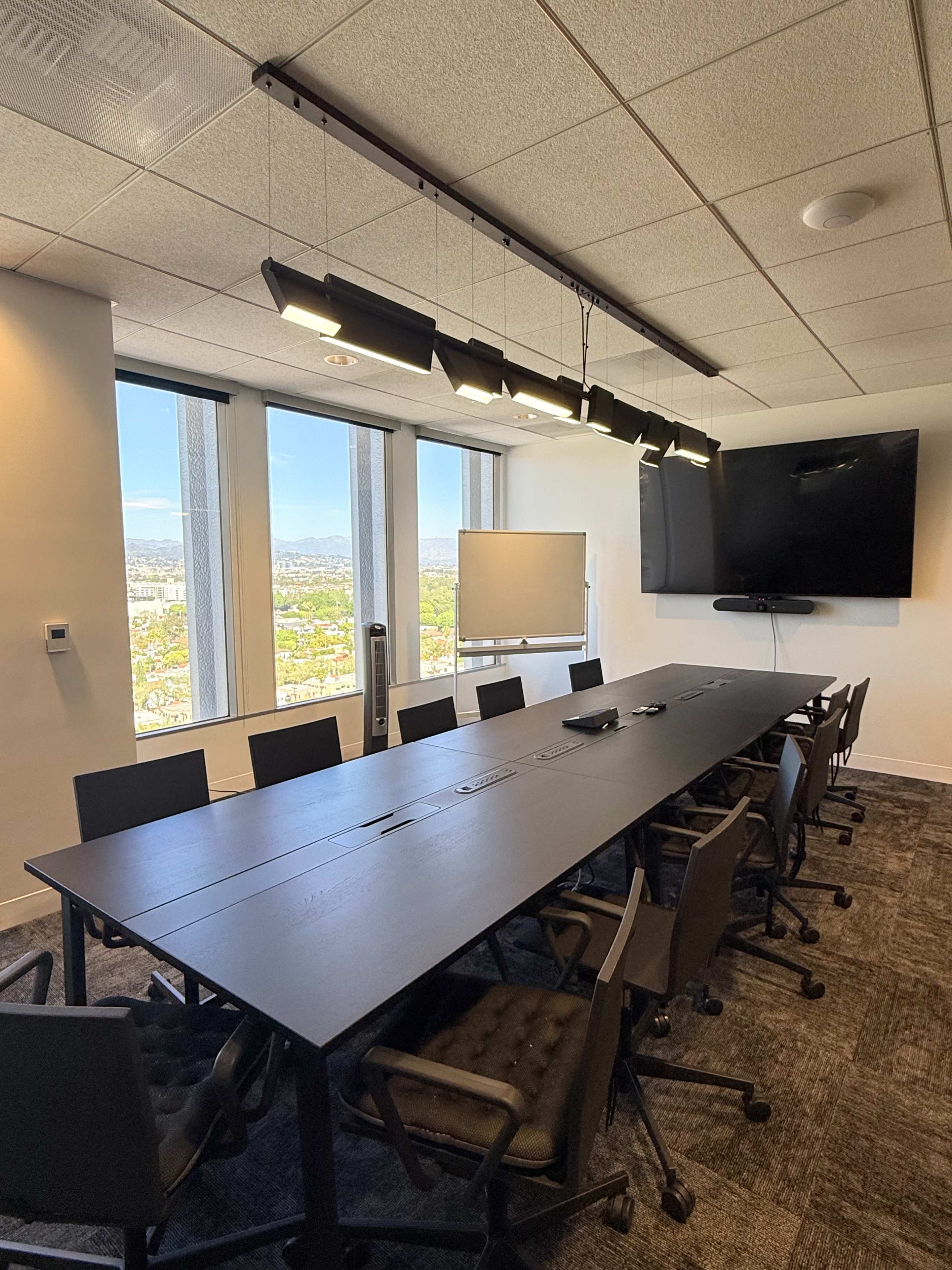 A rectangular conference room features a long table surrounded by chairs, large windows offering a city view, and a wall-mounted screen.