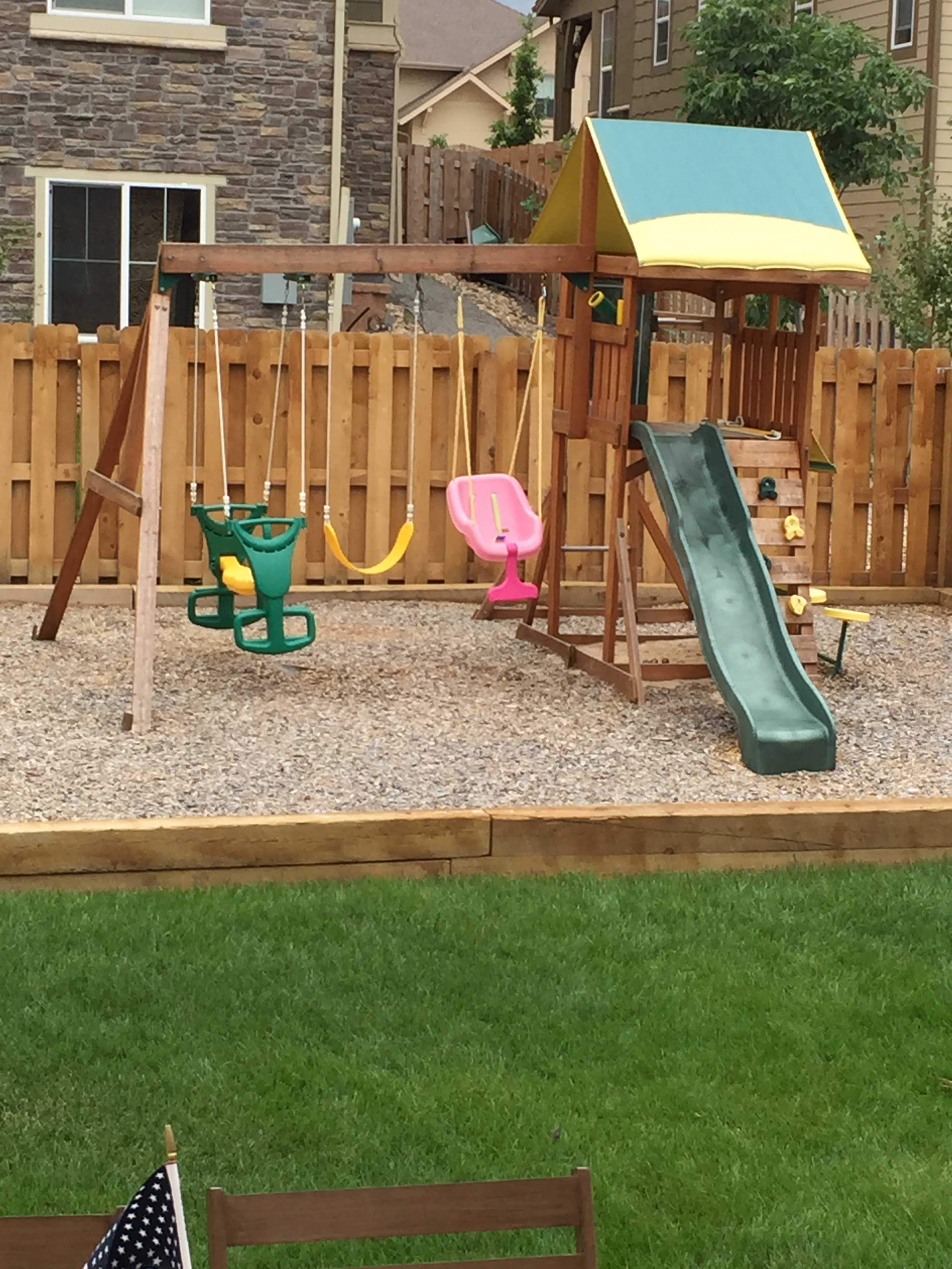 A wooden playground structure with a slide, swings, and a climbing area, surrounded by gravel and a wooden fence.