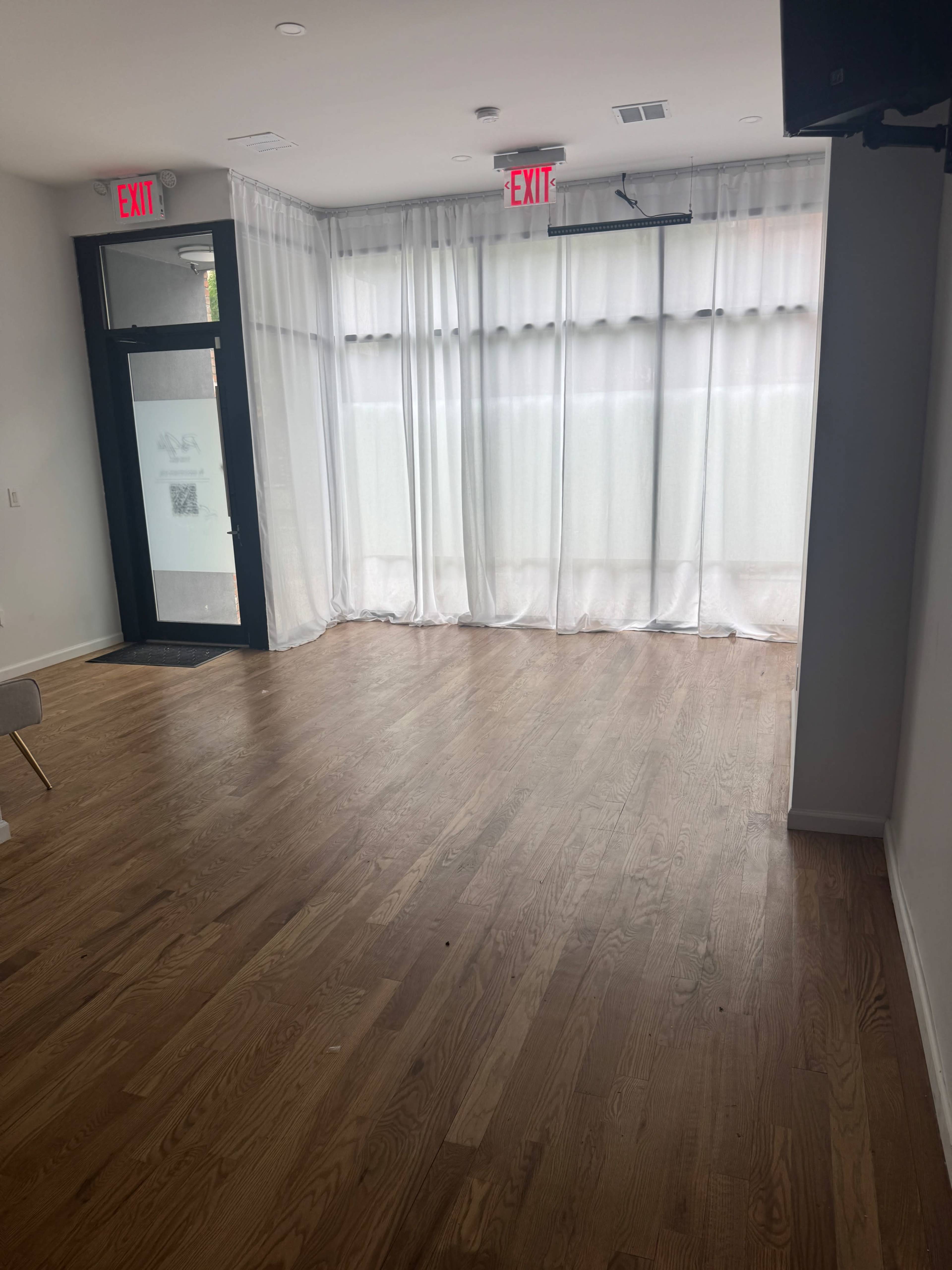 The room features a wooden floor, large windows covered with sheer curtains, and two exit signs above a doorway.