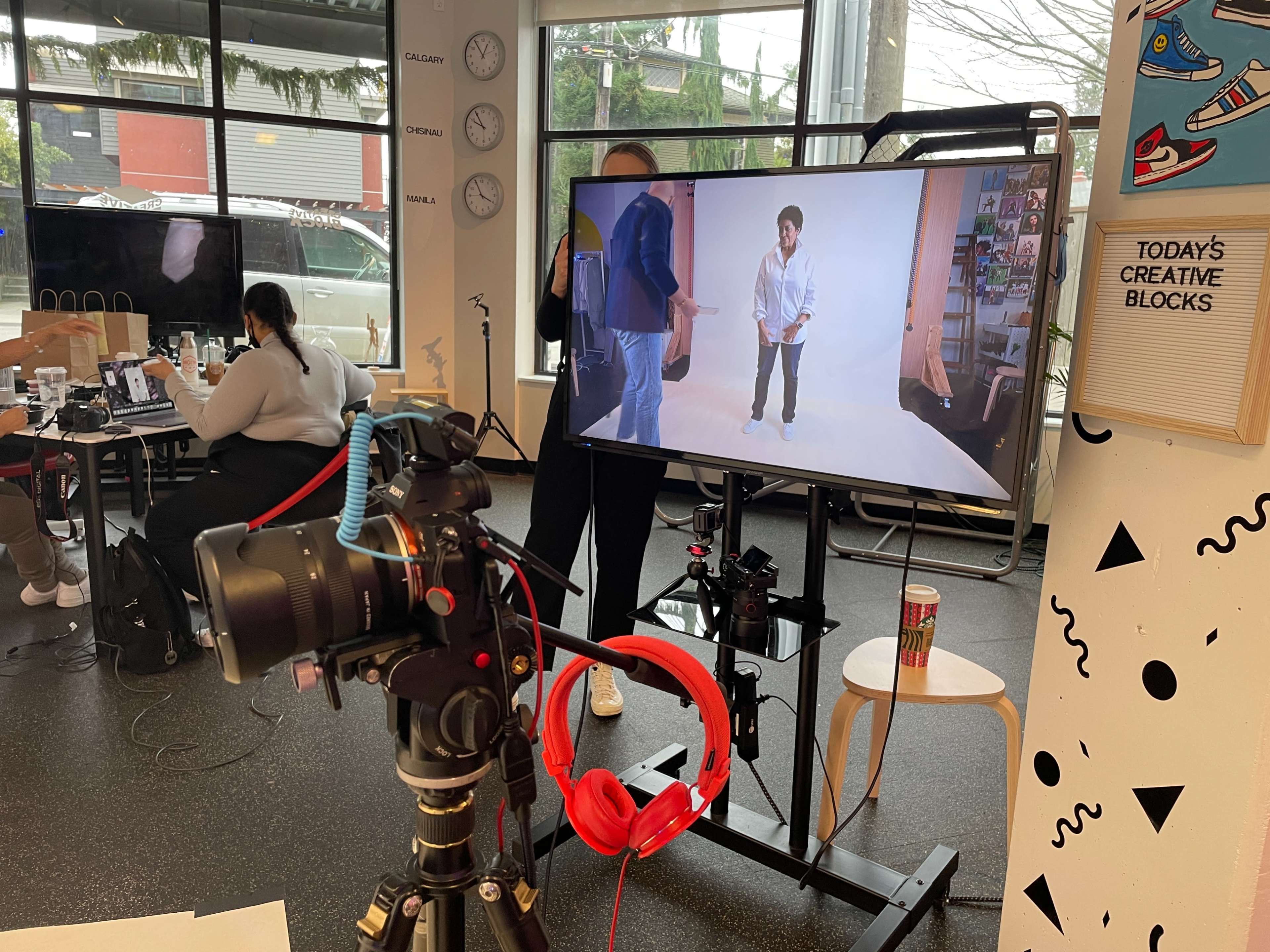 A camera on a tripod is positioned in front of a television screen displaying a person posing, while people work at tables in the background.