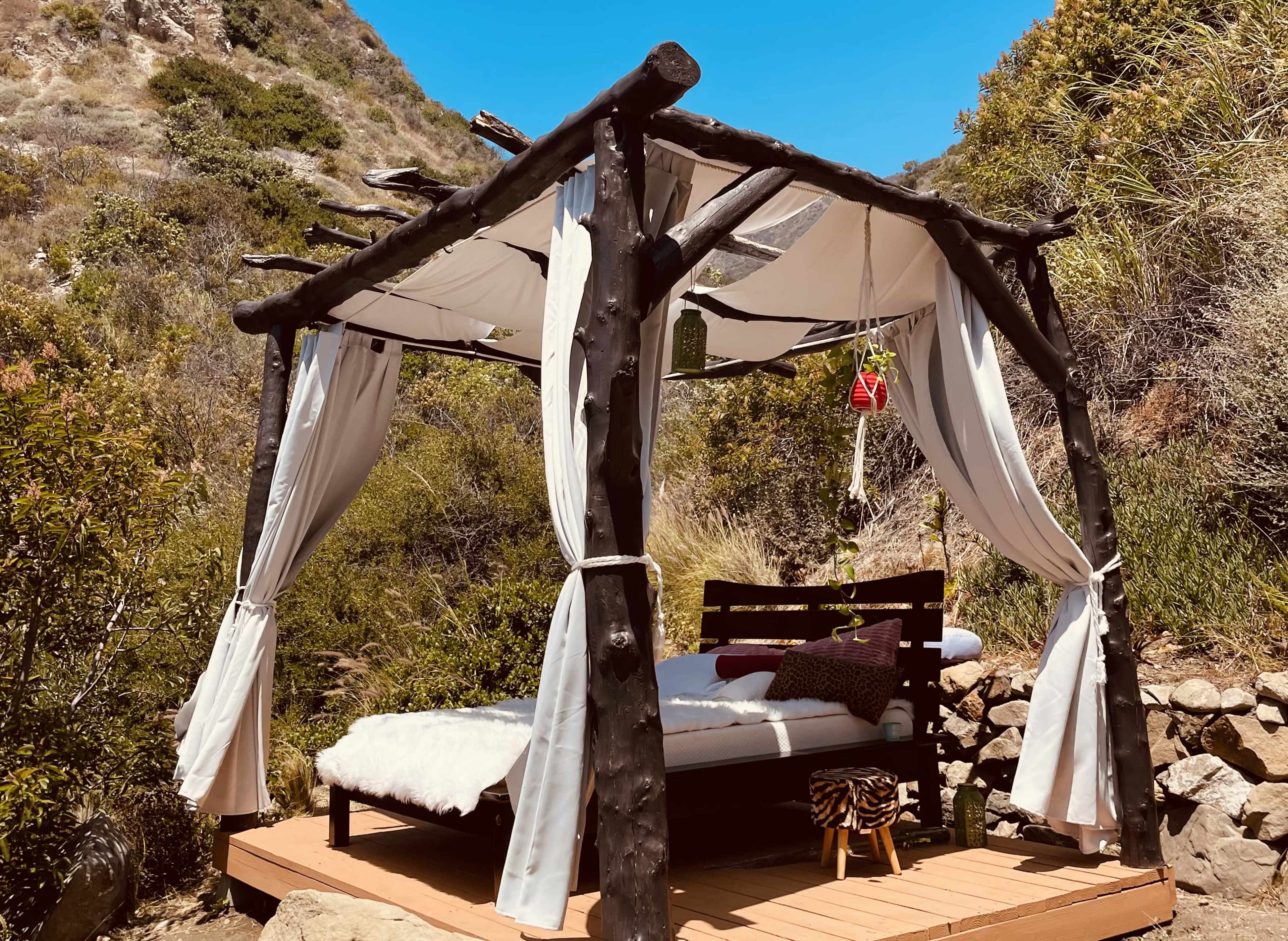 A wooden canopy bed with white curtains is set on a wooden platform surrounded by greenery and rocky terrain.