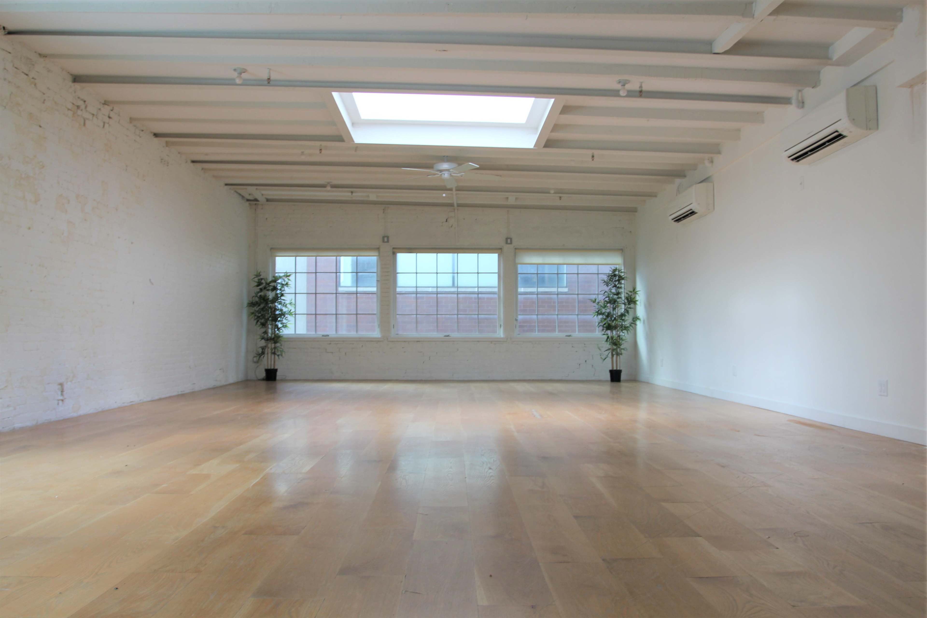 The image shows a spacious room with wooden flooring, white brick walls, a skylight, and two large windows with potted plants in the corners.