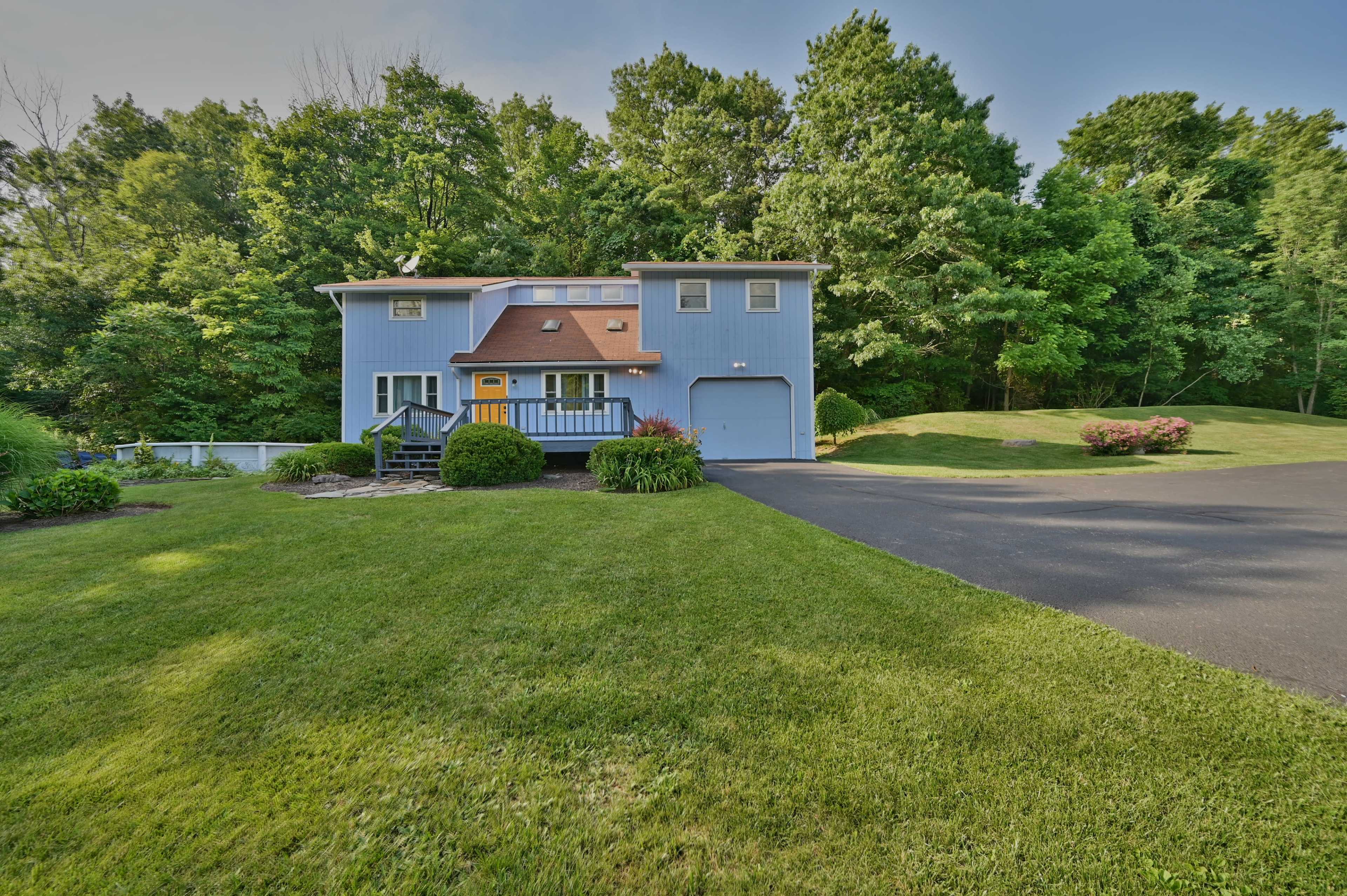 A two-story blue house with a brown roof sits on a green lawn, surrounded by trees and a paved driveway.