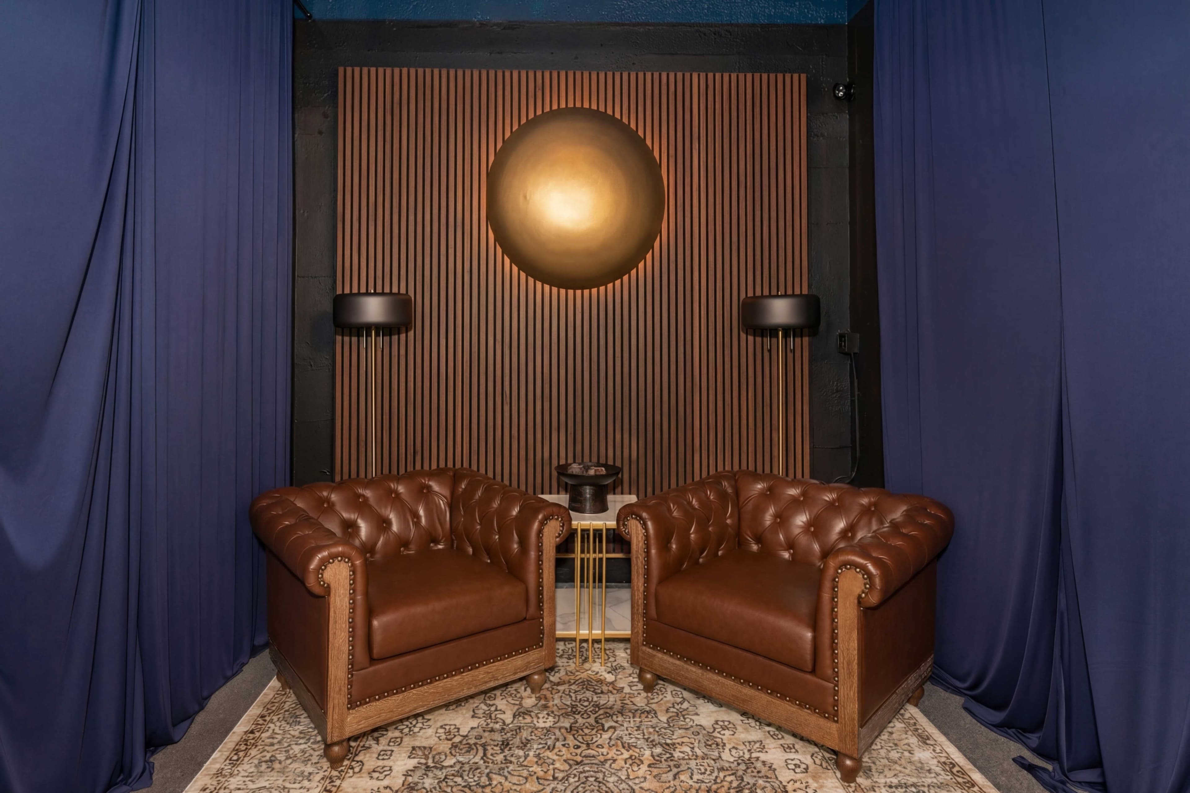 Two dark brown leather armchairs are positioned facing each other in front of a wooden slat wall, with a circular gold accent above them and a small table in between.