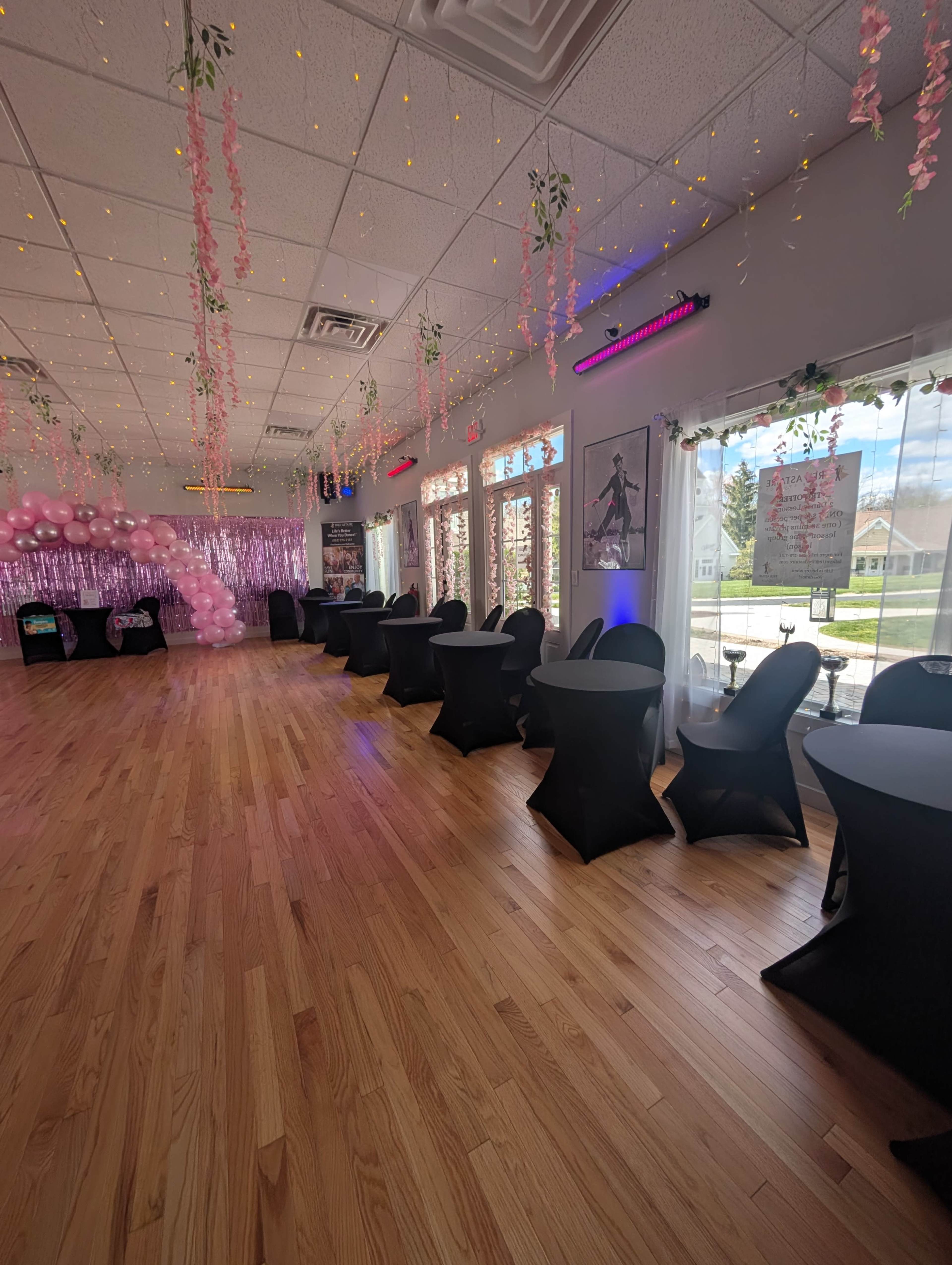 The interior of a decorated event space features tables covered with black cloth, pink balloons, and strings of lights hanging from the ceiling.