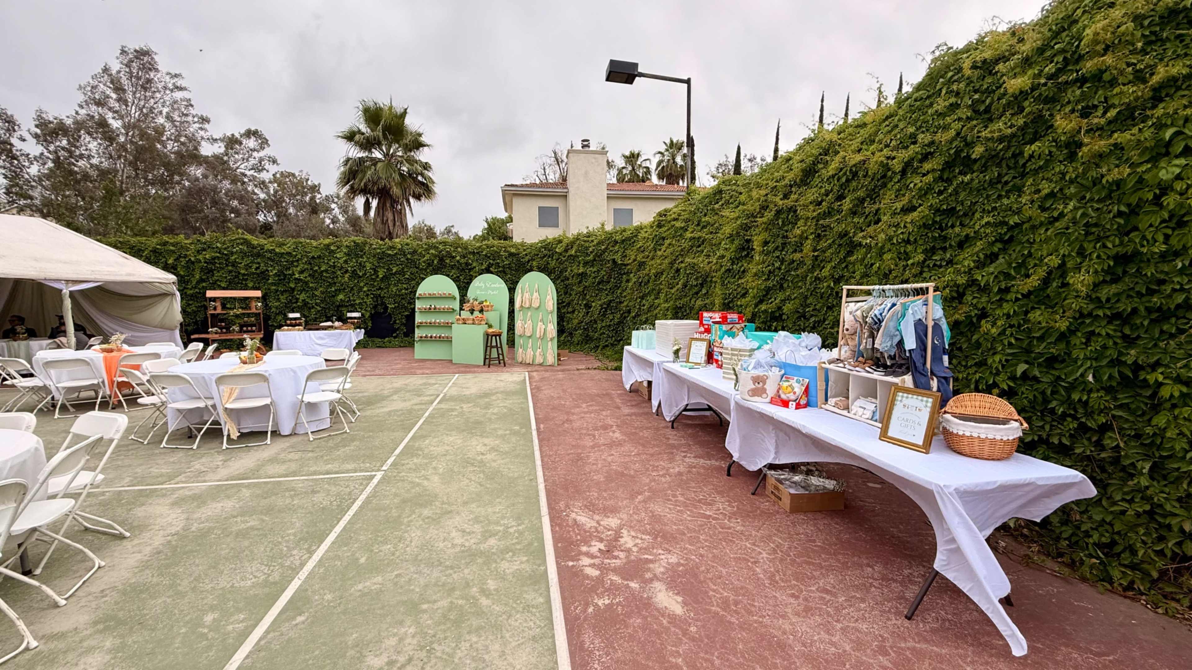 The outdoor space features white tables set up for an event, surrounded by a lush green hedge, with decorated display tables on one side showcasing various items.