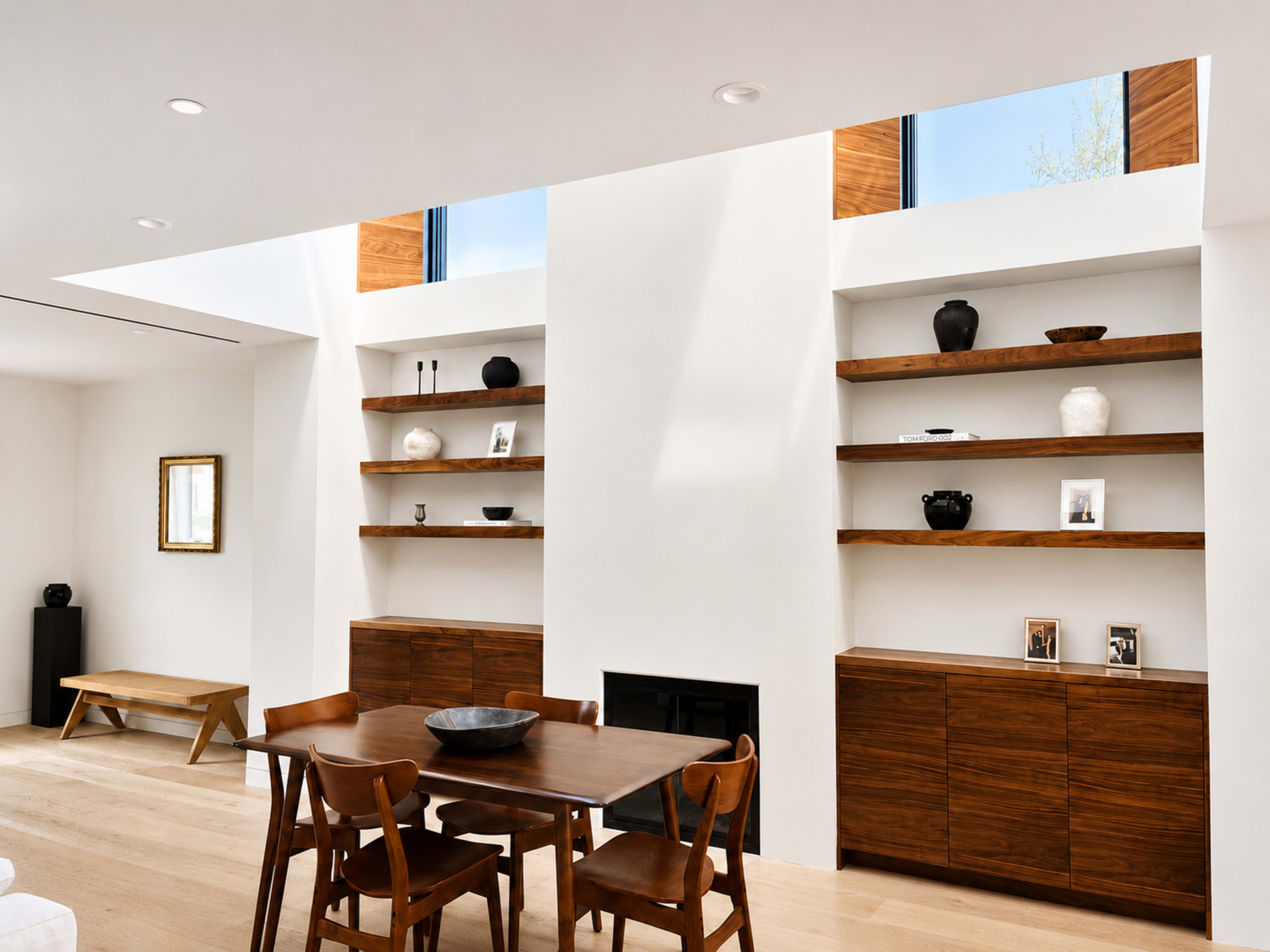 The image shows a modern living space featuring a wooden dining table with chairs, flanked by shelves filled with decorative items and photographs, under skylights that illuminate the room.