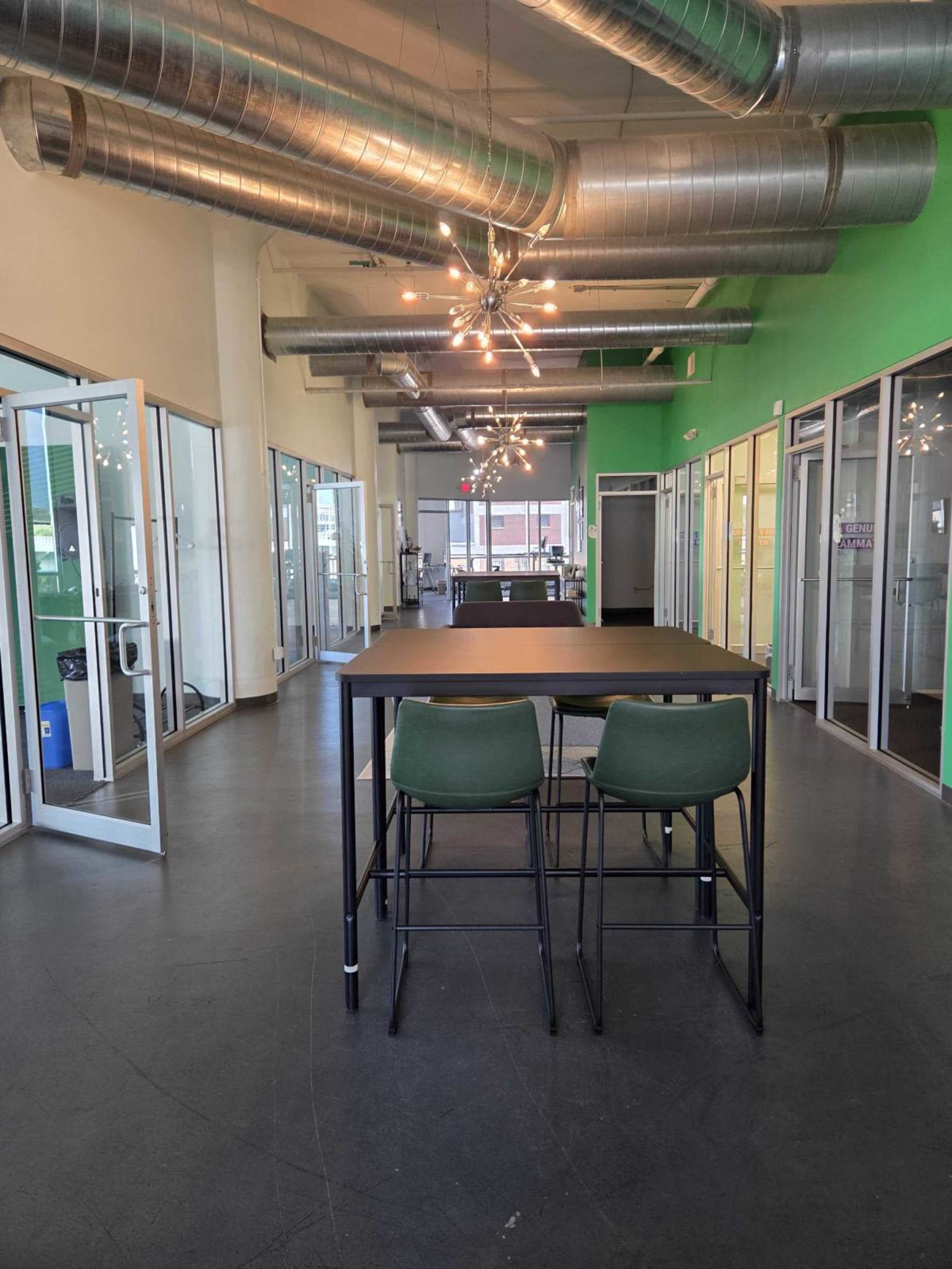 The image shows a modern office space with a central table and chairs, surrounded by glass-walled meeting rooms and green accent walls.