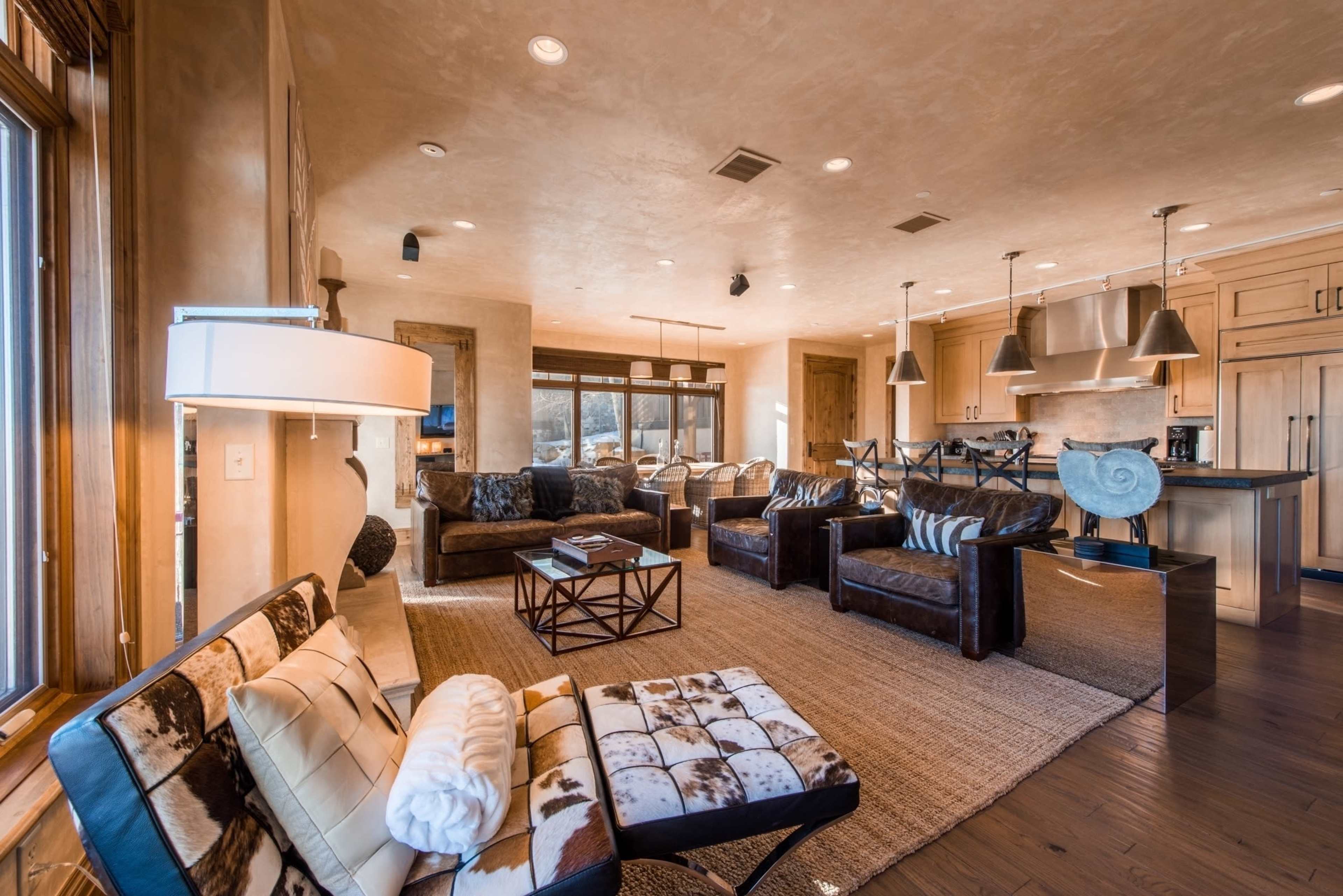 A modern living room with a combination of leather seating, large windows, and a kitchen area in the background.