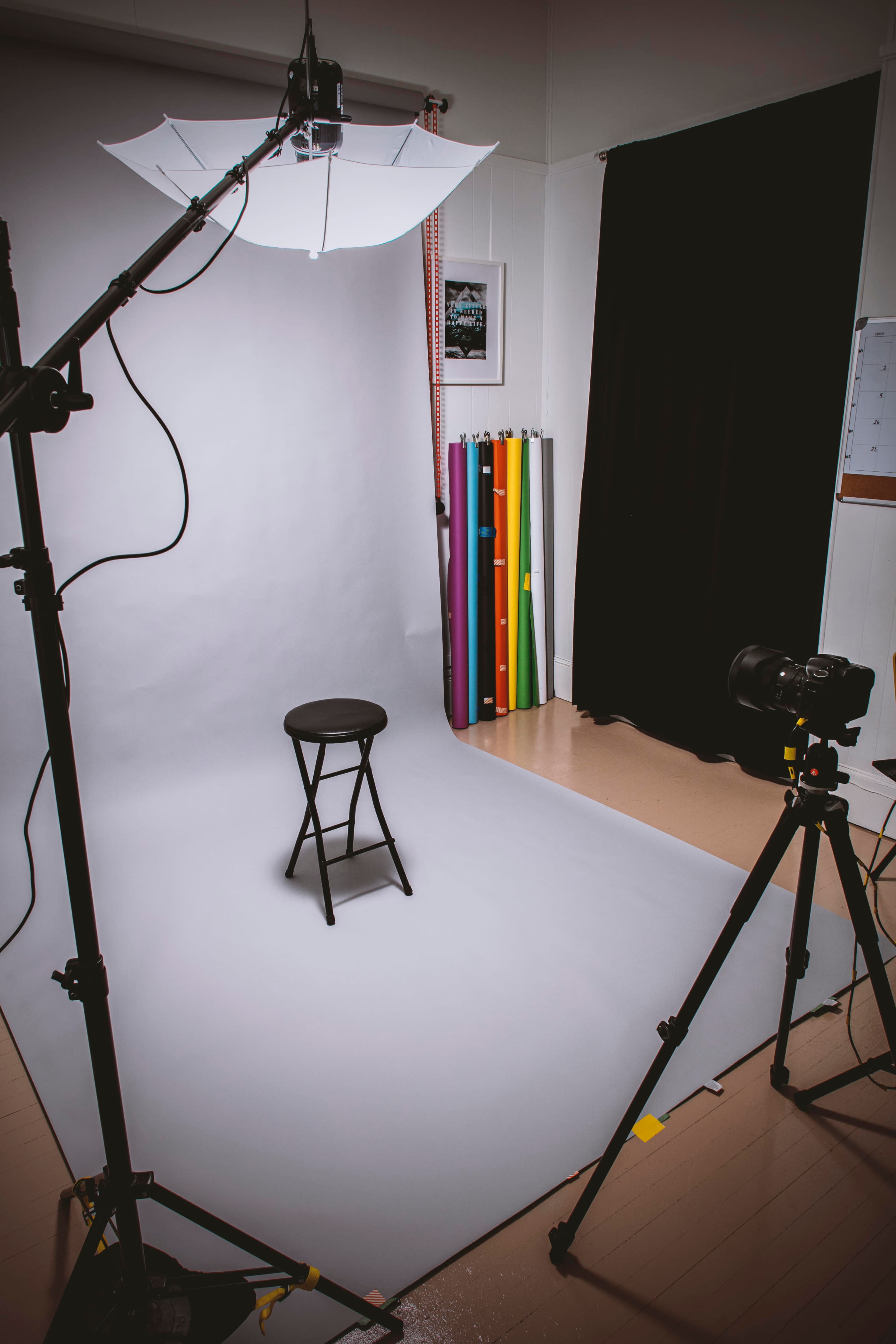 The image shows a photography studio setup with a black stool positioned on a gray backdrop, studio lights overhead, and a collection of colorful backdrop rolls in the background.