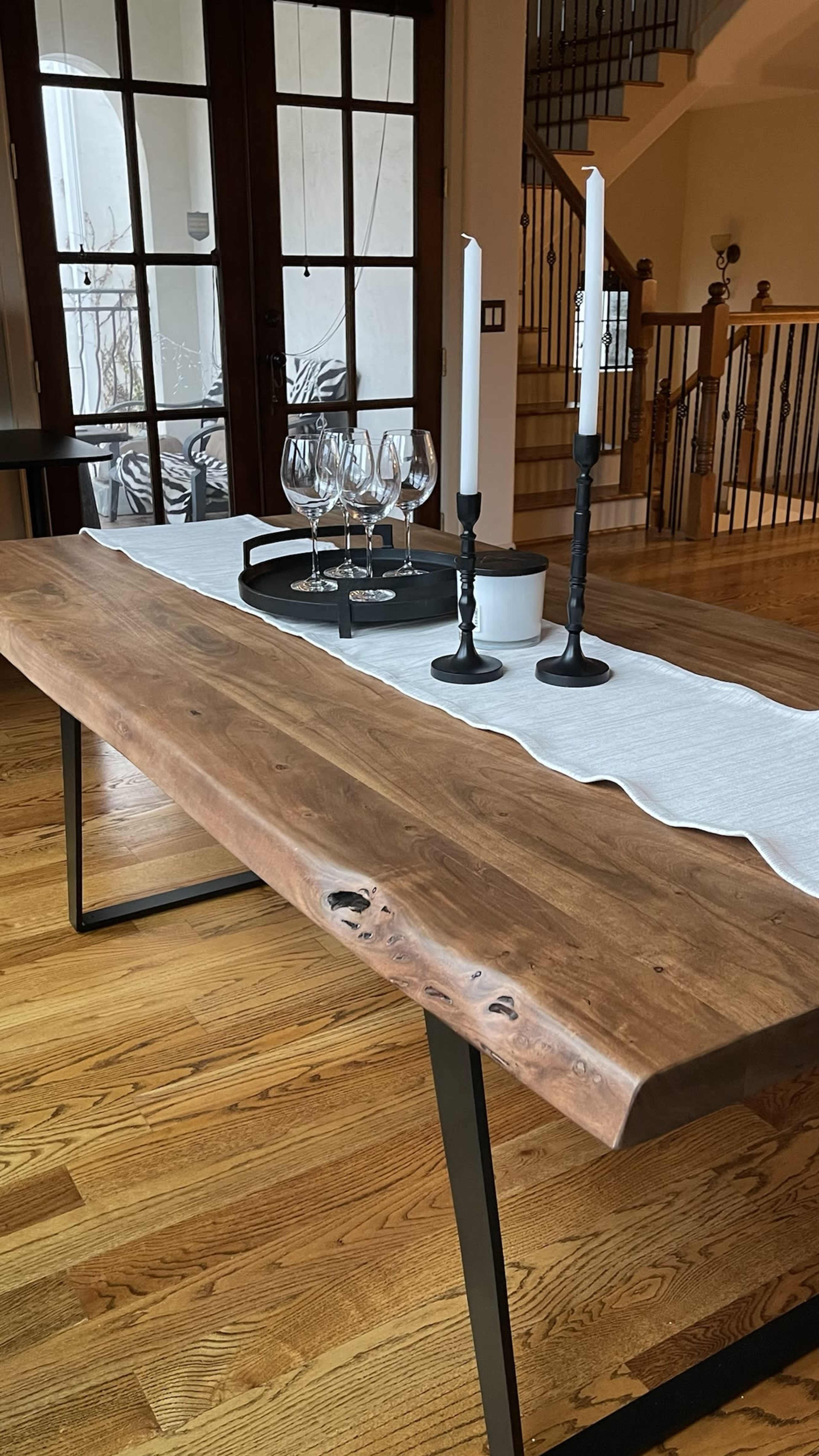 A wooden dining table with a natural edge is set with a white runner, two candles, a bowl, and a tray of glasses.