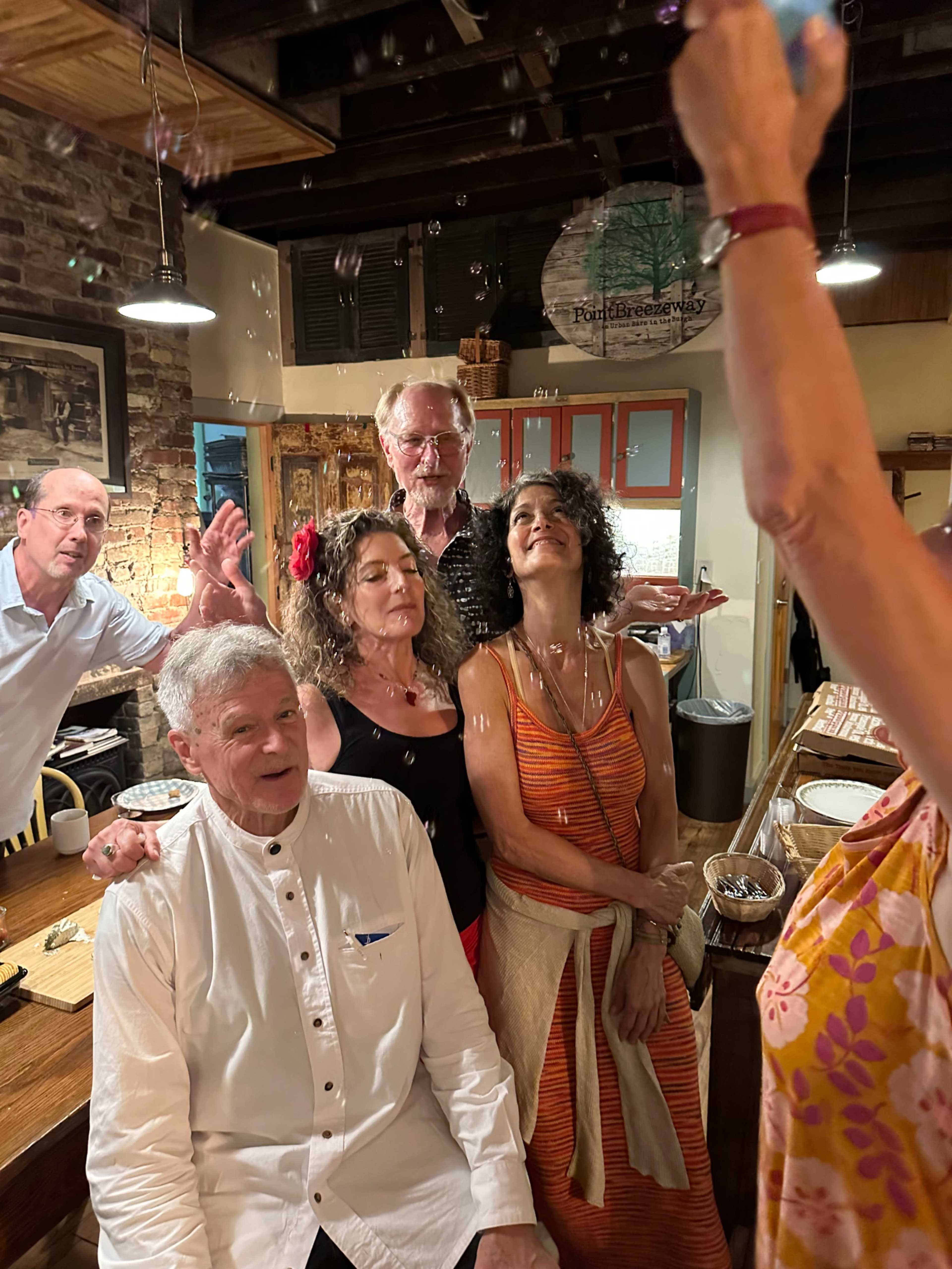 A group of people stands around a seated man as bubbles float in the air during a gathering in a rustic kitchen.