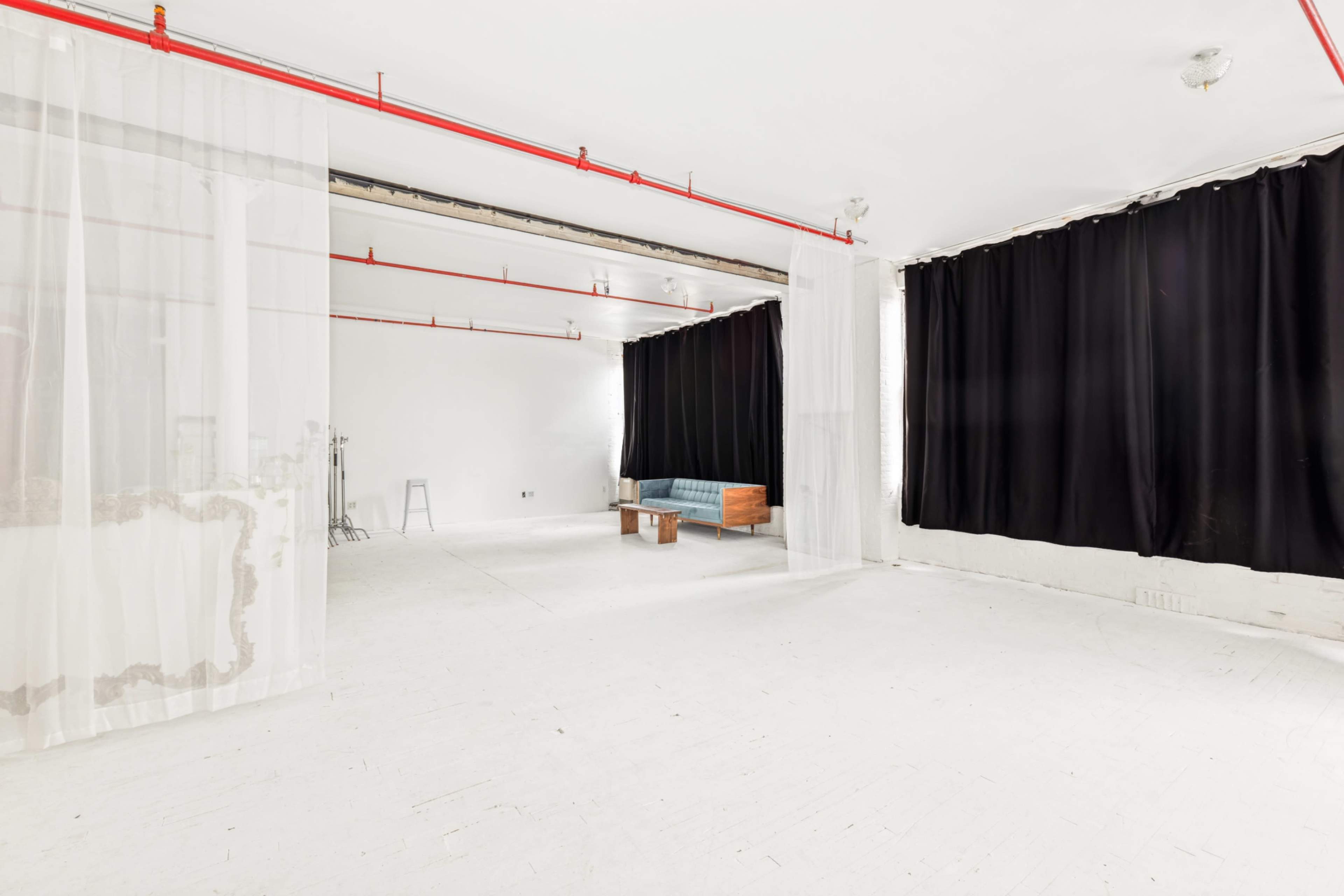 The image shows a spacious, empty interior room with white walls and flooring, featuring large black curtains and a wooden bench.