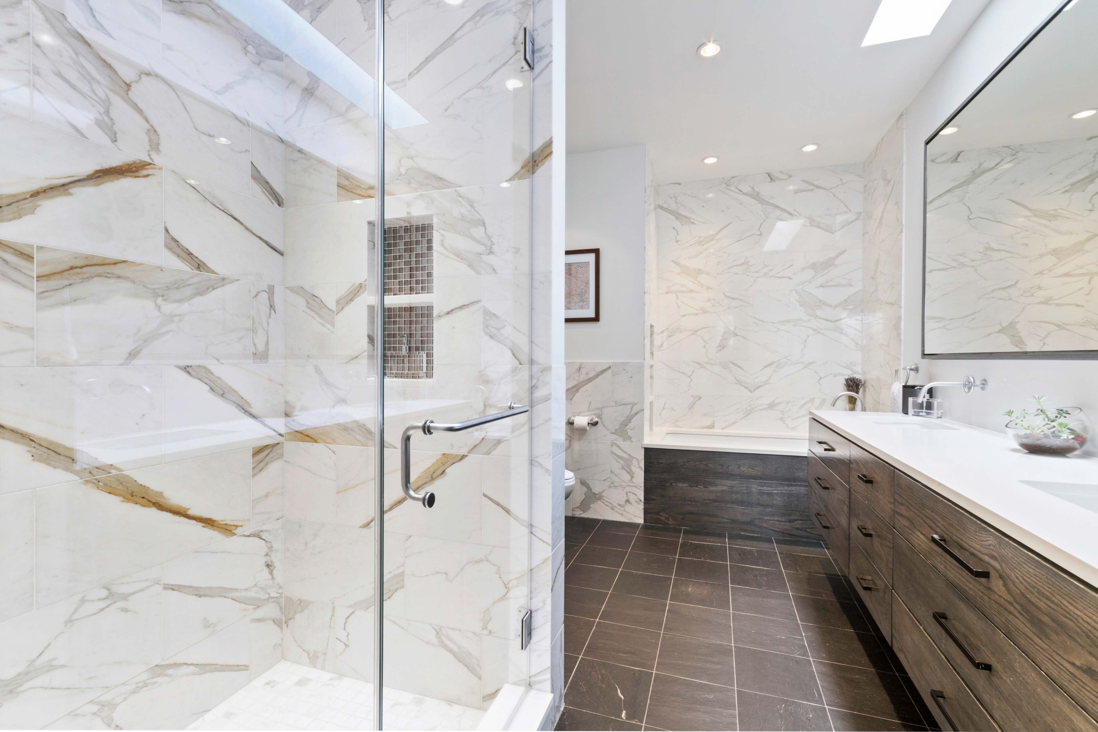 A modern bathroom featuring a glass shower, marble walls, and a double vanity with wooden cabinetry.