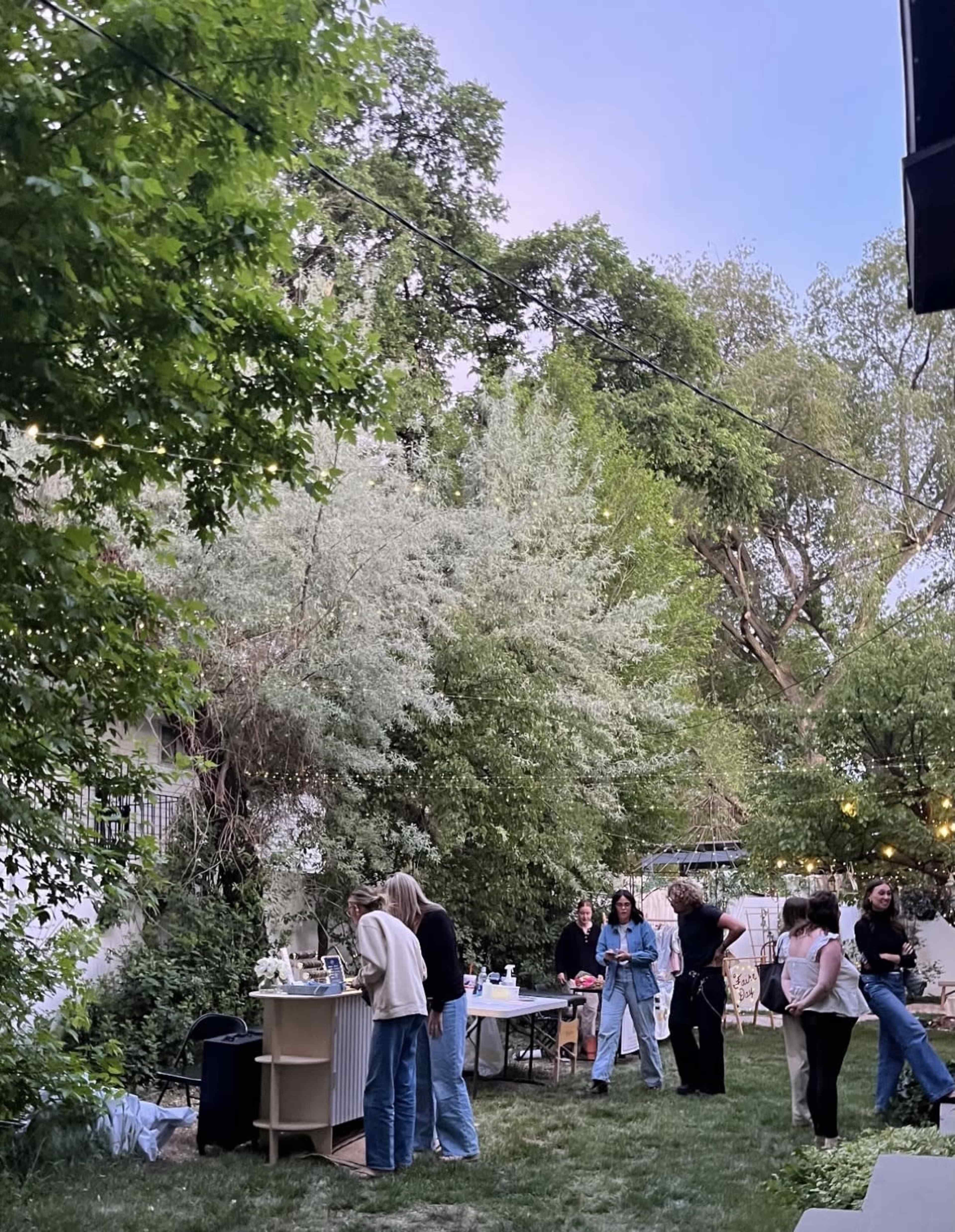 A group of people socializes in a backyard surrounded by trees, with tables set up for refreshments.