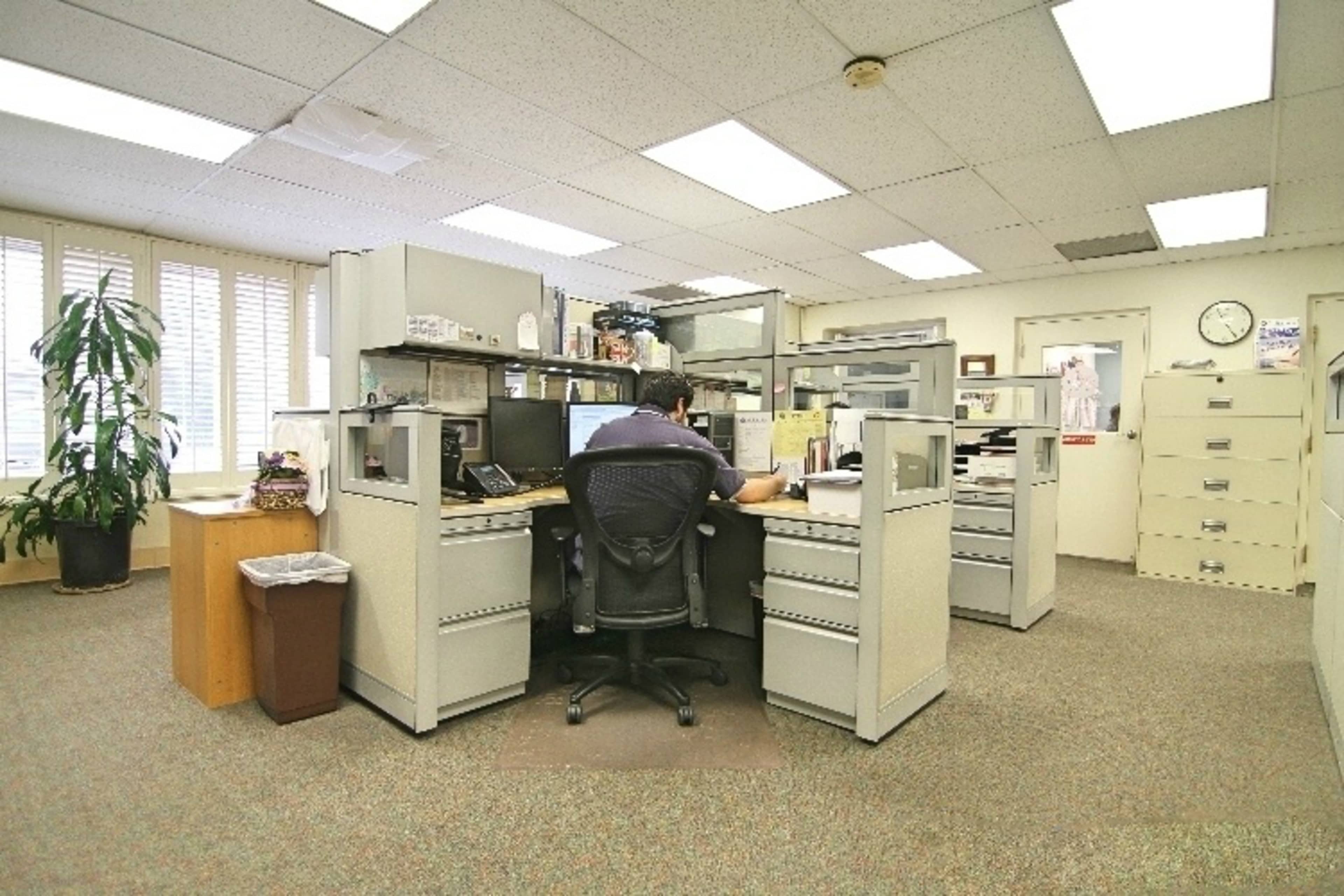 An employee sits at a cubicle desk with filing cabinets and office equipment in a well-lit office space.