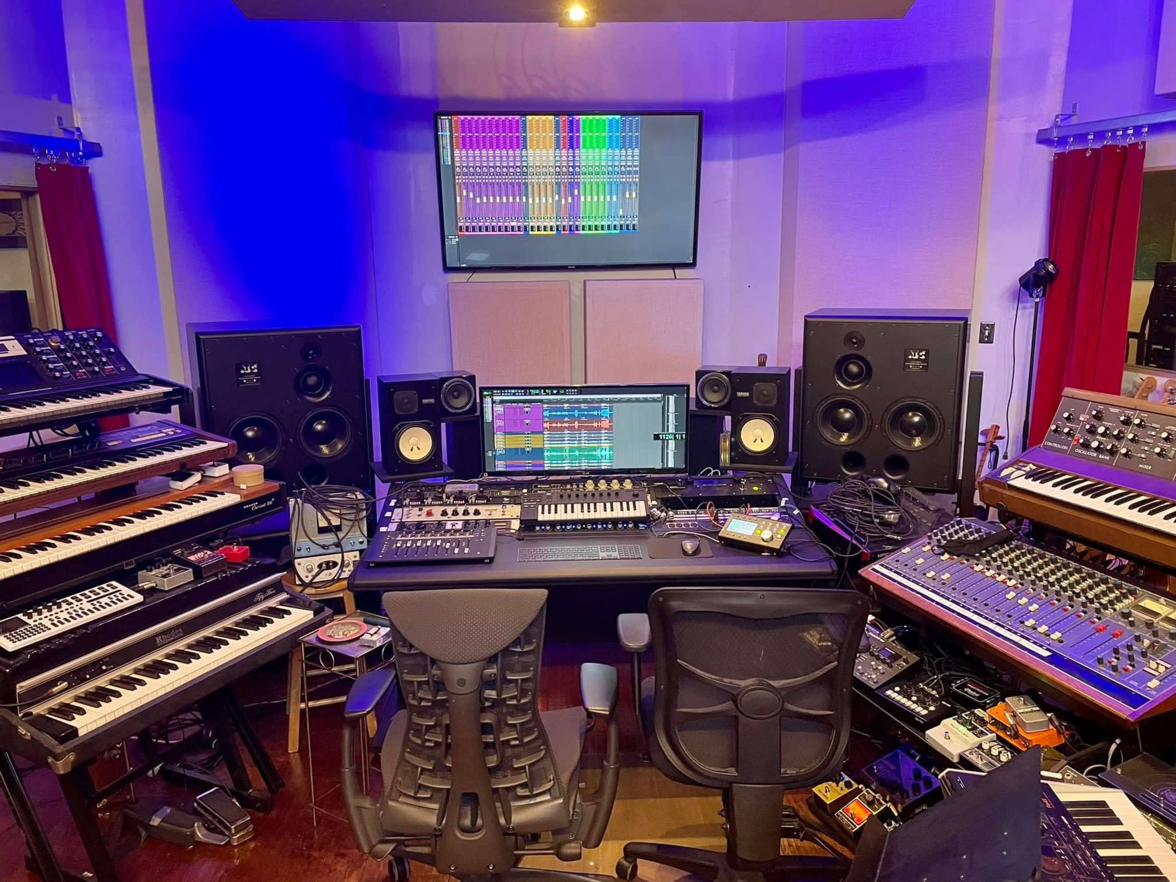 The image shows a music studio with various keyboards, synthesizers, monitors, and a central mixing console surrounded by speakers.