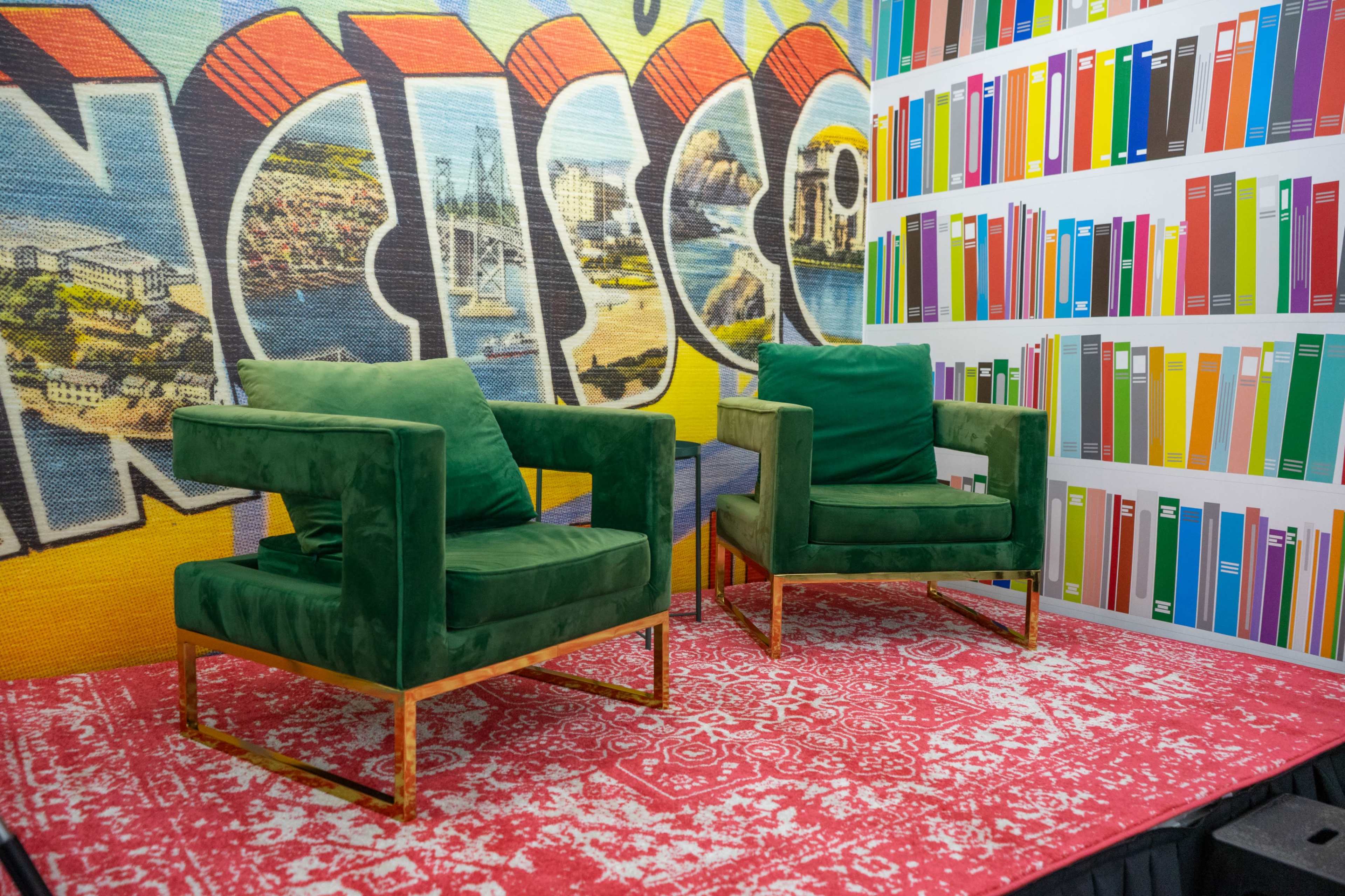 The image shows two green velvet chairs on a patterned red carpet in front of a colorful mural and a wall decorated with illustrations of books.