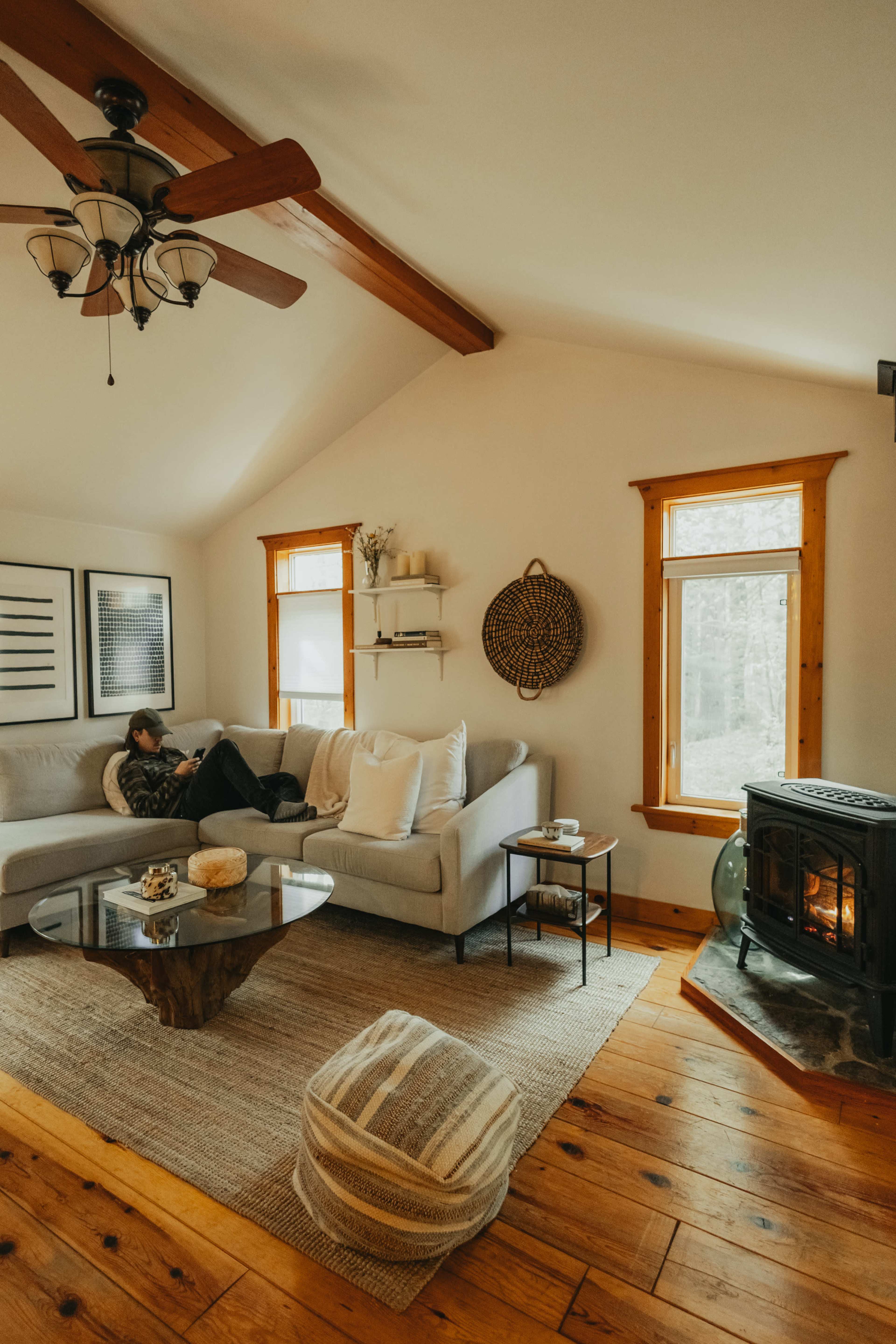 A person sits on a light gray sofa in a cozy living room with wood accents and a small stove, surrounded by large windows and a coffee table.
