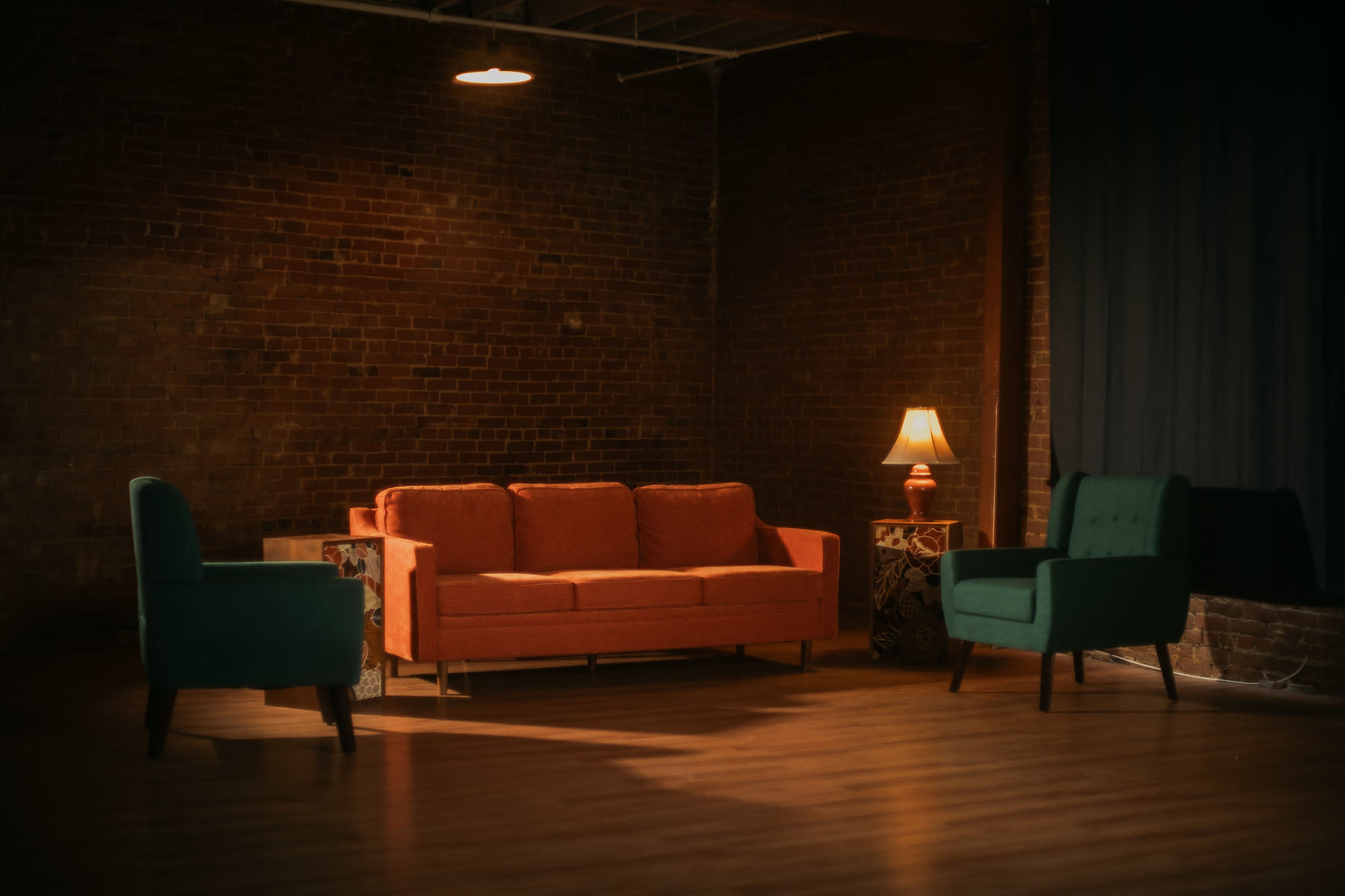 The image shows a minimalist lounge area featuring an orange couch, two green chairs, and a lamp, set against a backdrop of exposed brick walls.