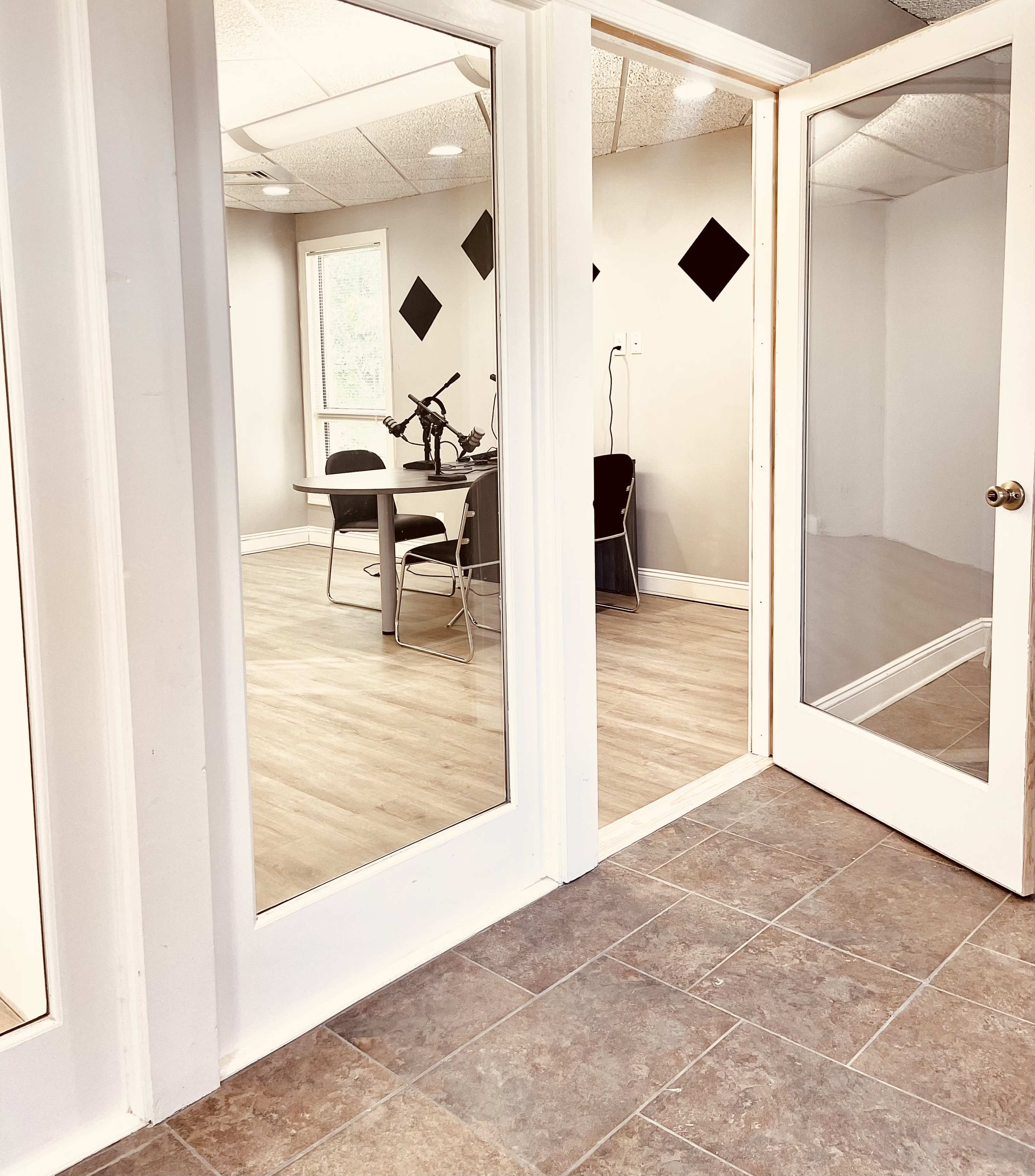 A corridor with two large glass-paneled doors leading into a conference room, featuring a table and chairs, against a backdrop of light-colored walls and a wooden floor.