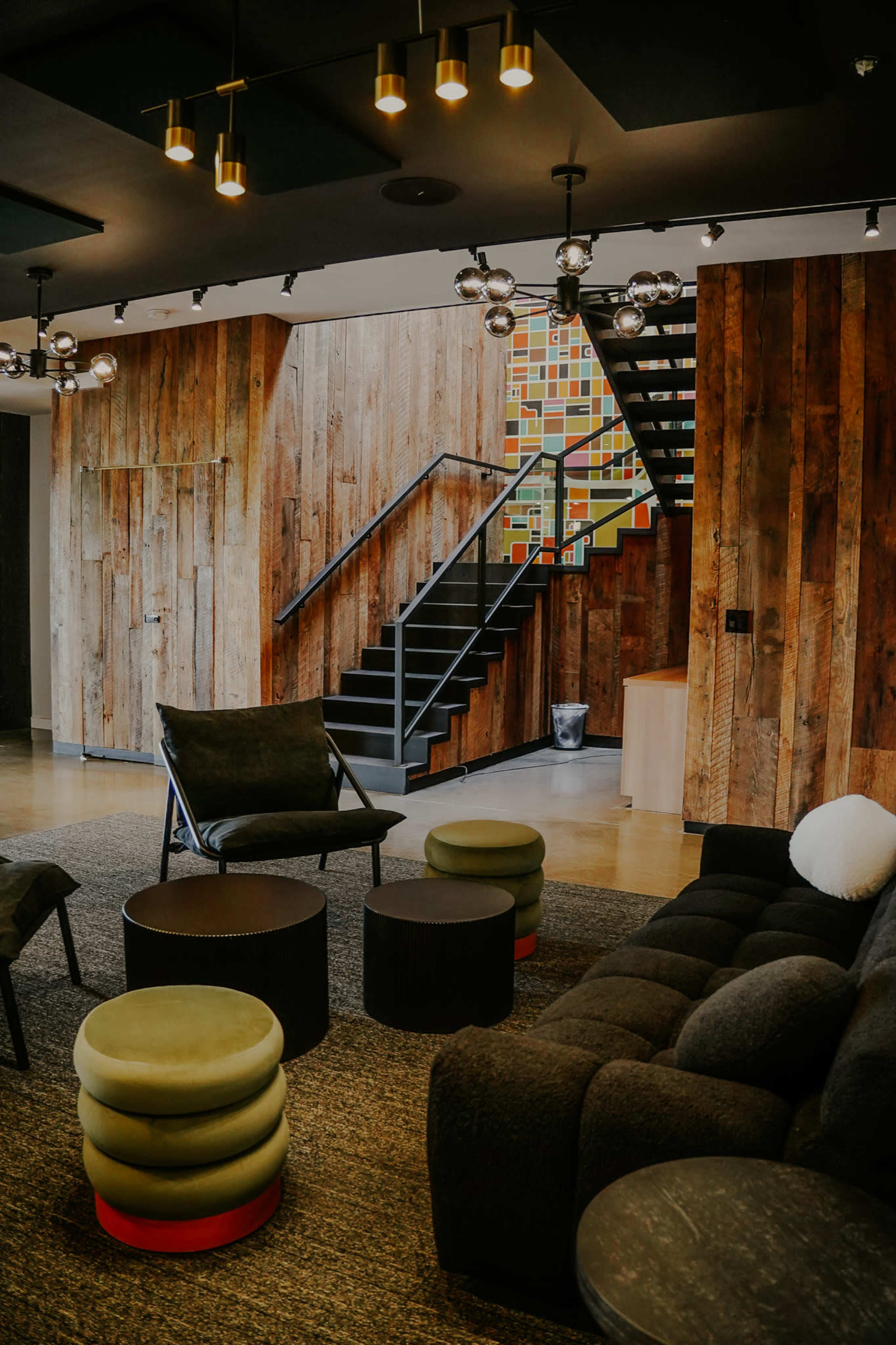The image shows a modern interior space featuring a wooden staircase, a seating area with circular stools, and textured walls.