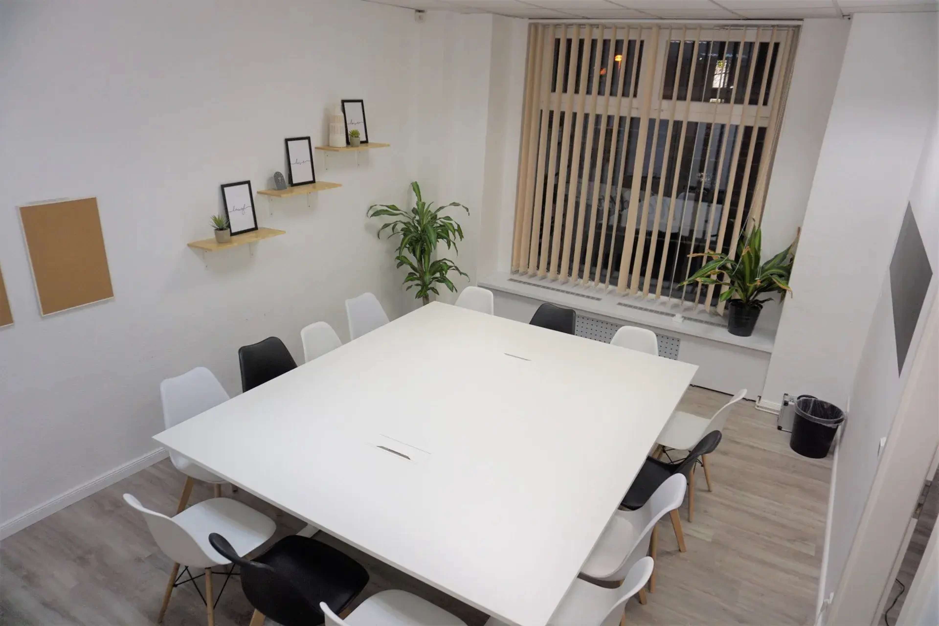 A spacious conference room features a large white table surrounded by chairs and is illuminated by natural light from a window.