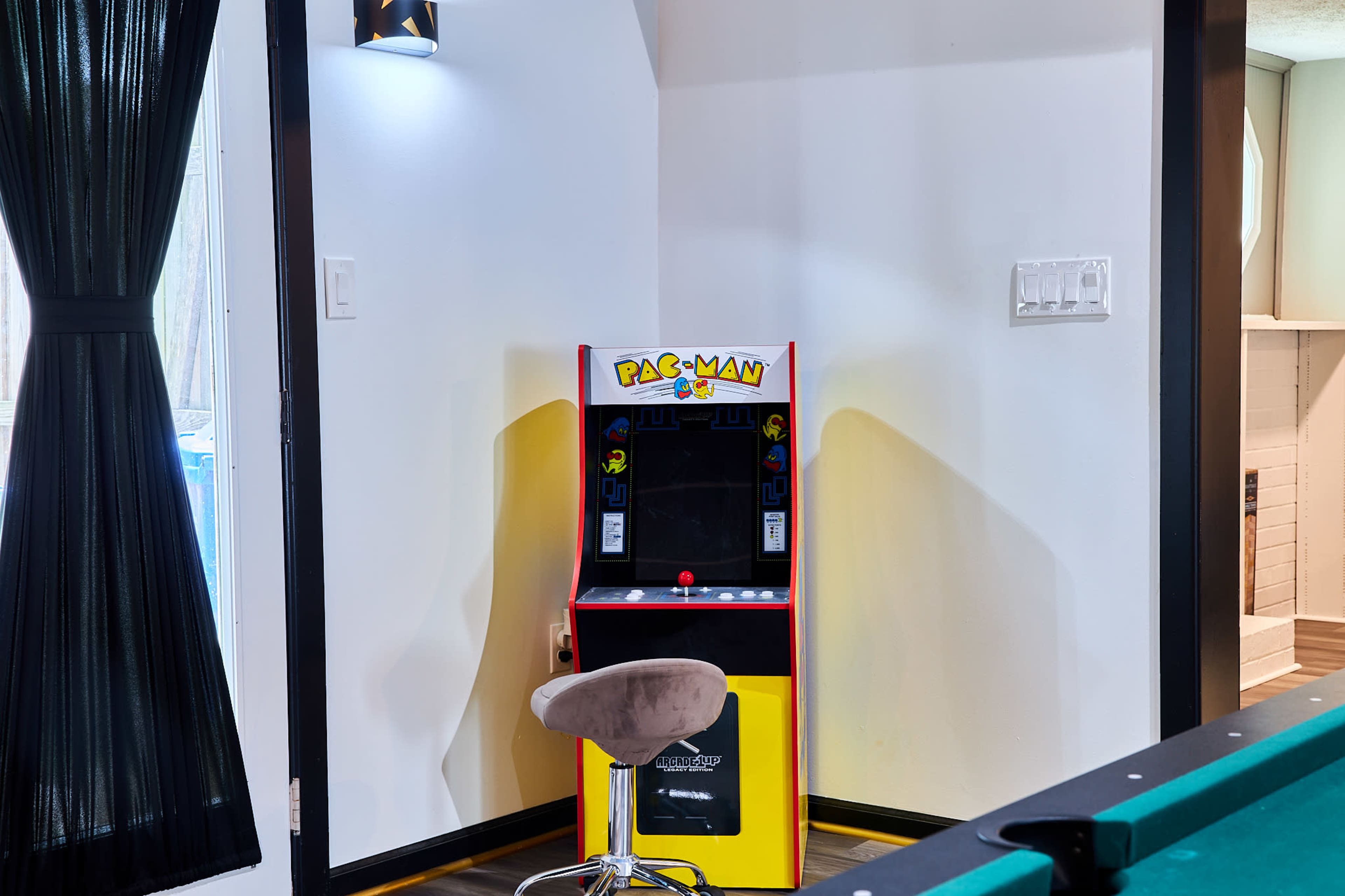 A vintage Pac-Man arcade machine is positioned against a white wall, accompanied by a gray stool.
