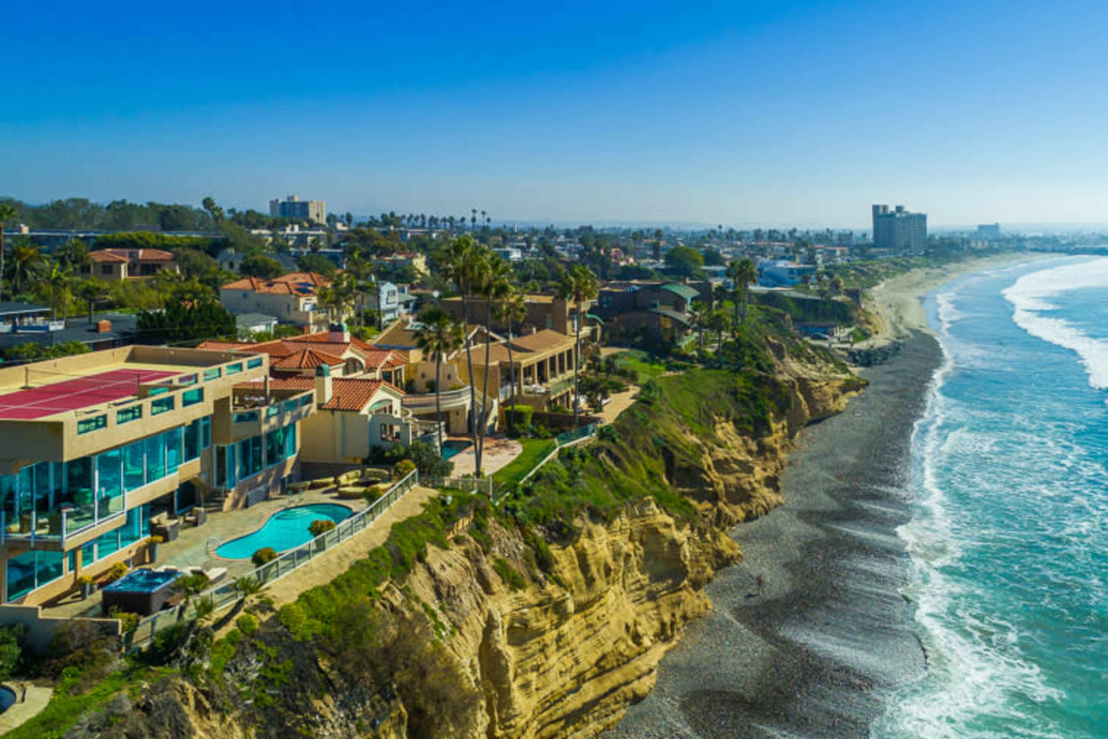 A coastal view features luxury homes perched along a cliff overlooking a sandy beach and ocean waves.