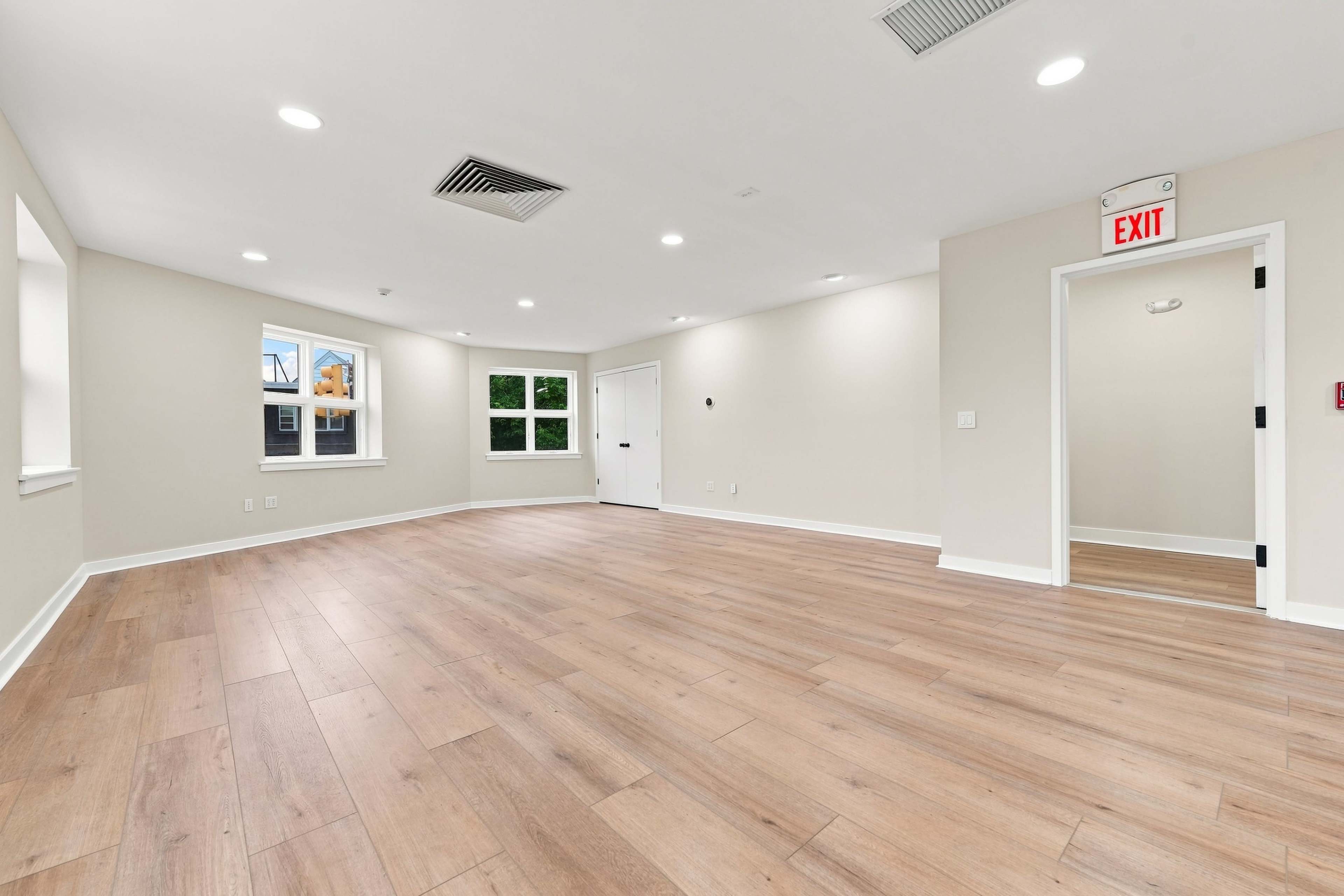 The image shows an empty room with wooden flooring, a door, and several windows allowing natural light.