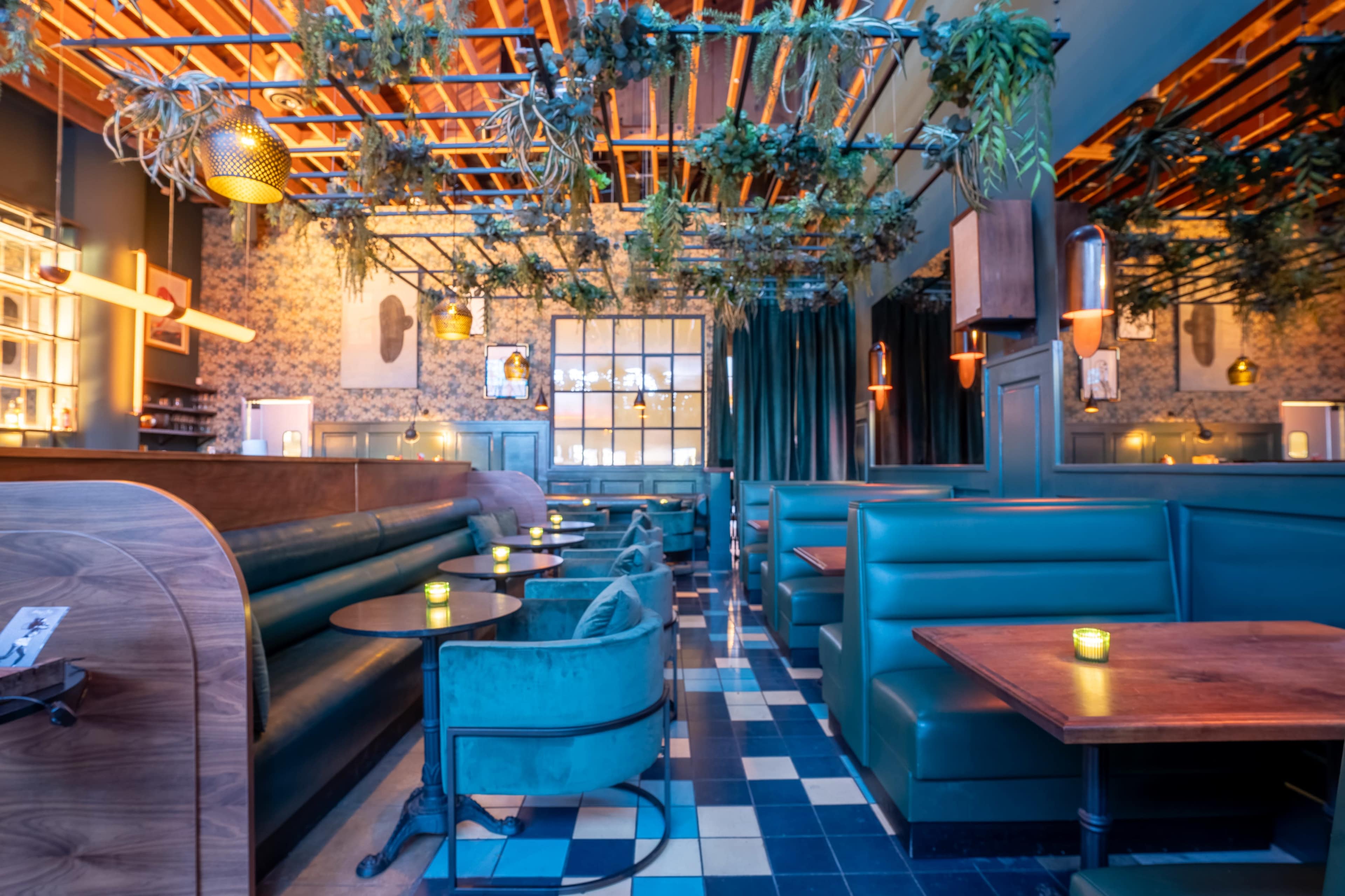 The image depicts a modern restaurant interior featuring green upholstered seating, wooden accents, and hanging plants, with tables arranged for dining.