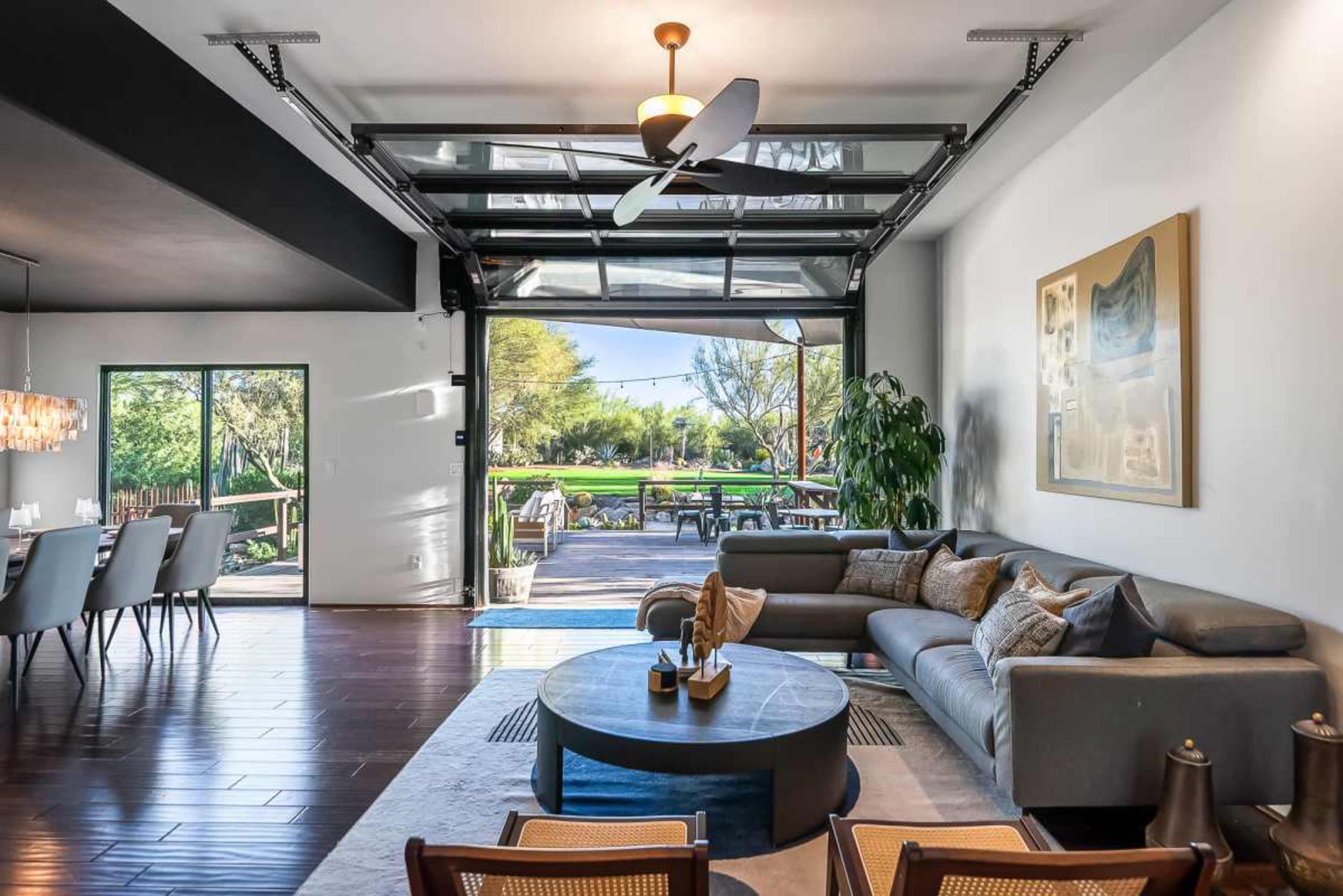The image shows a contemporary living space with a large sectional sofa, a coffee table, and a garage door that opens to a landscaped outdoor area.