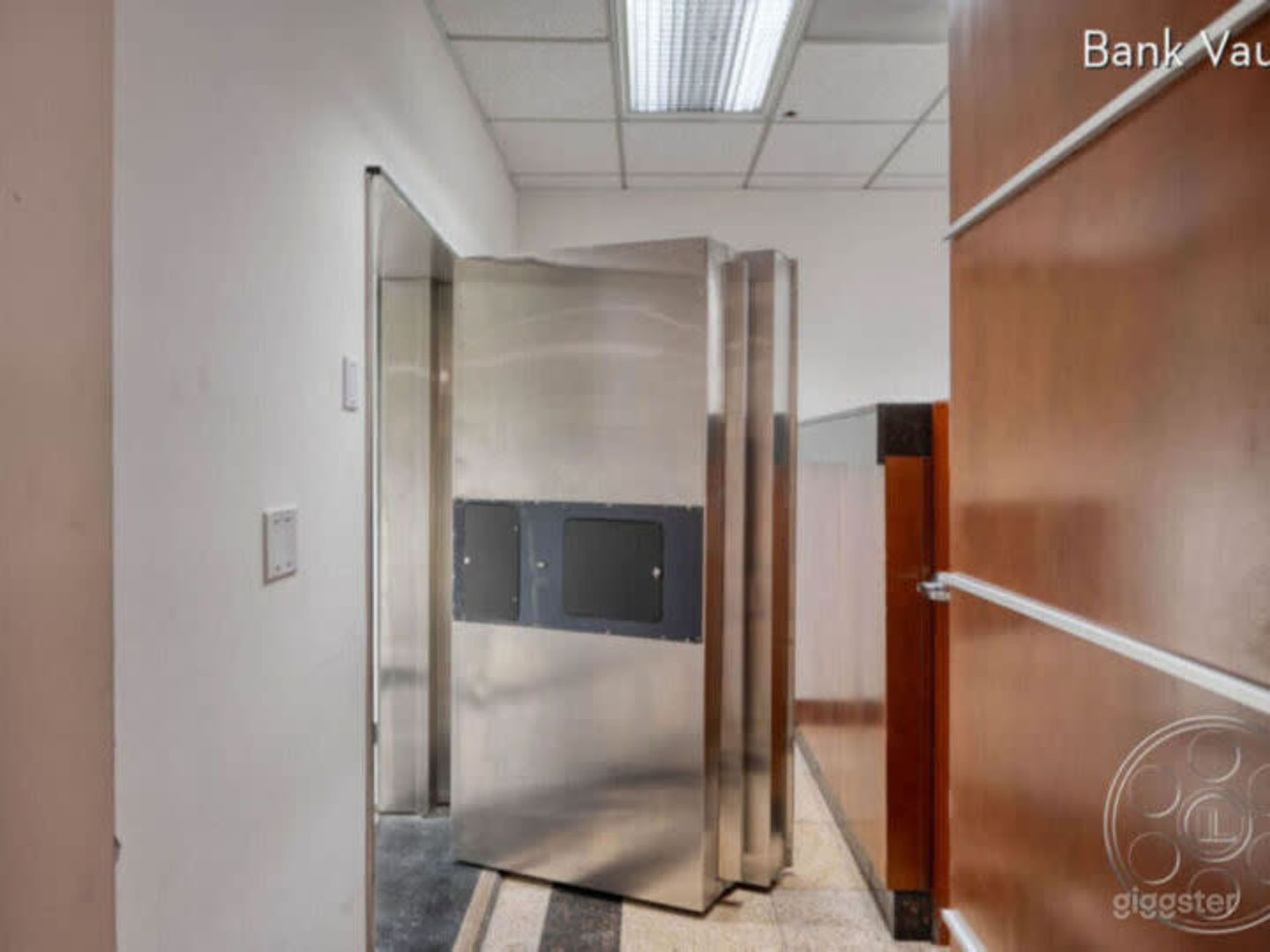 A stainless steel bank vault door stands partially open in a hallway next to wood-paneled walls.