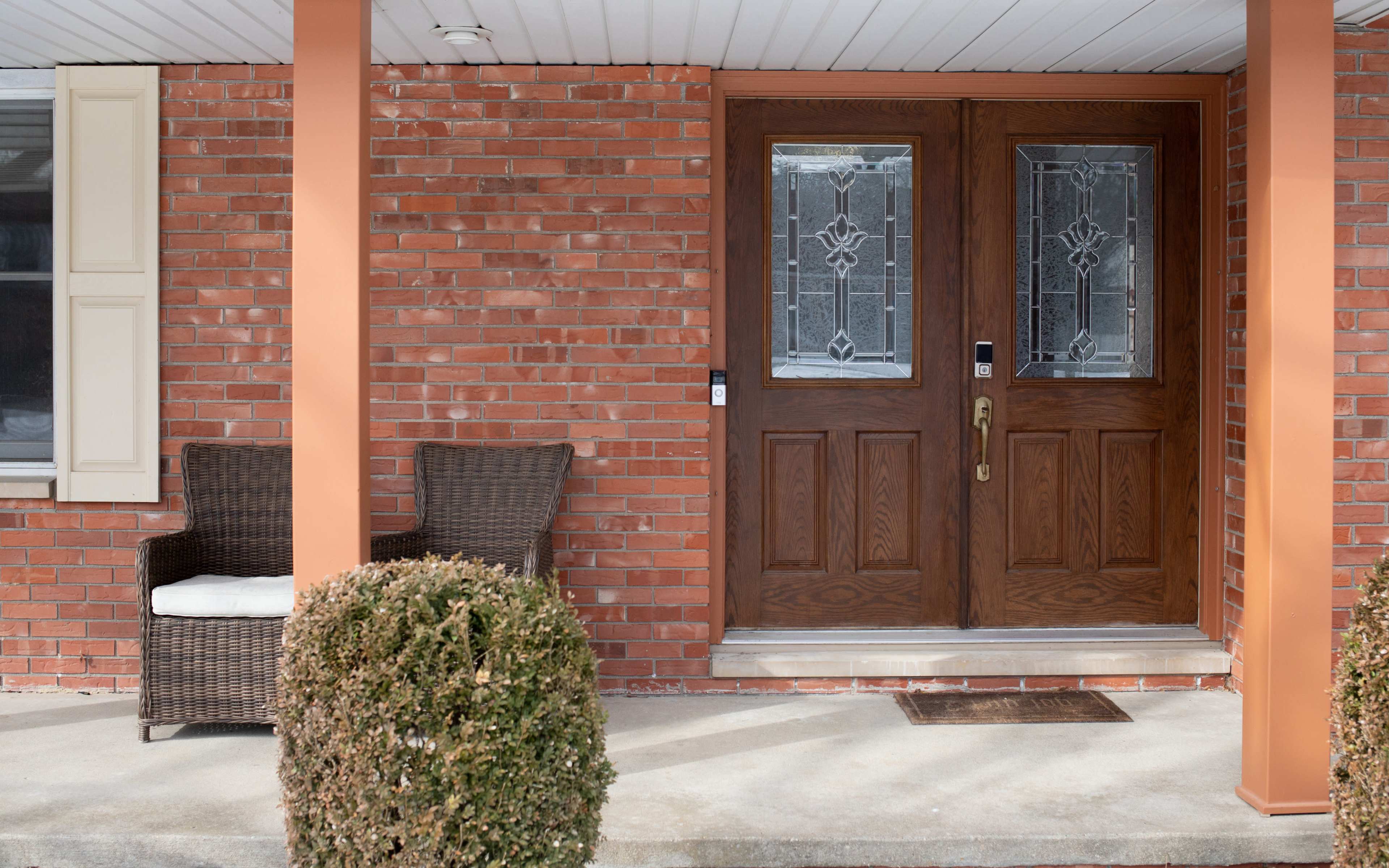 The image shows a front entrance with double wooden doors and a brick facade, flanked by a wicker chair and two neatly trimmed bushes.