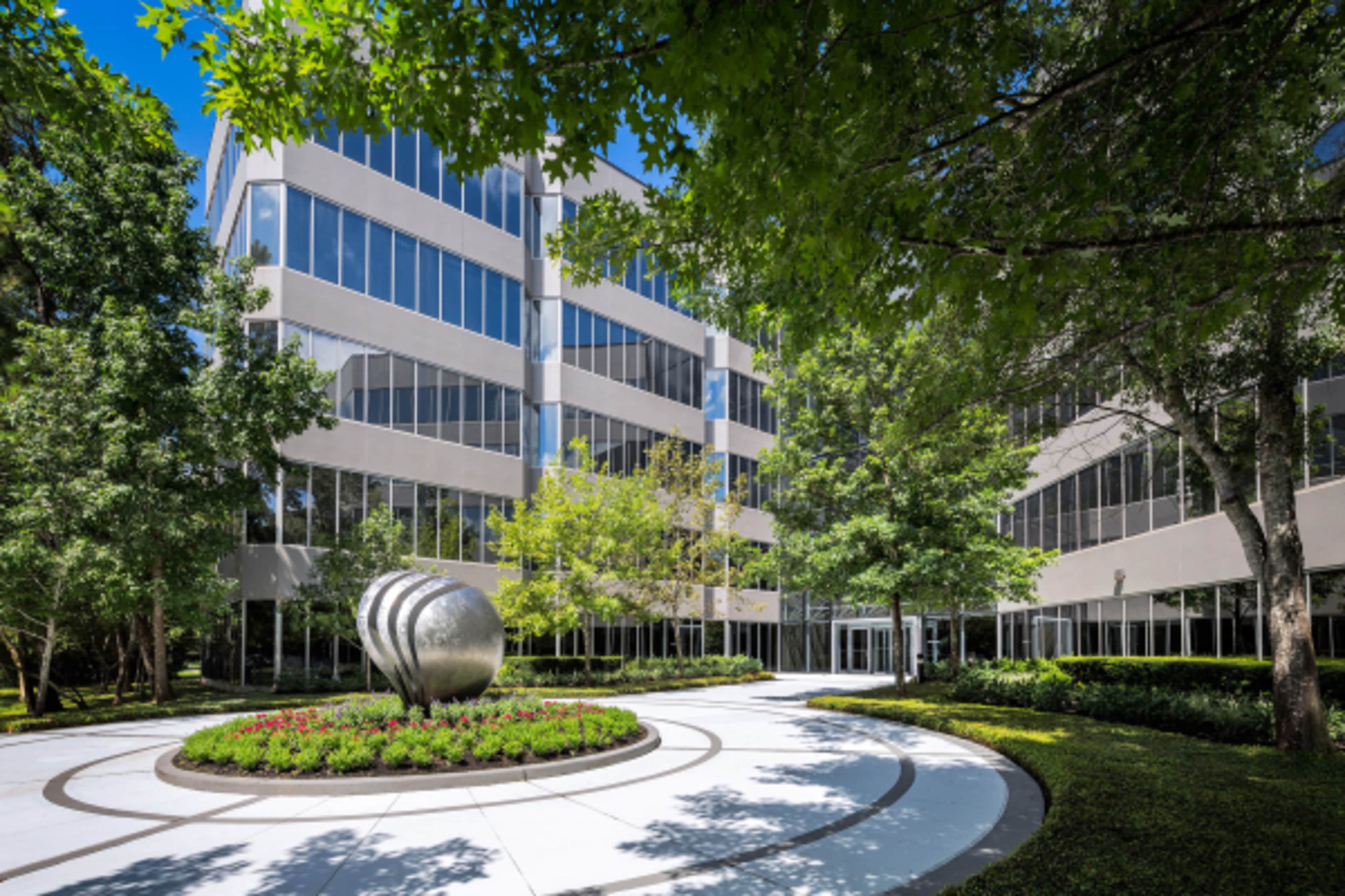 A modern office building is surrounded by landscaped greens and features a circular pathway with a decorative sculpture in the center.
